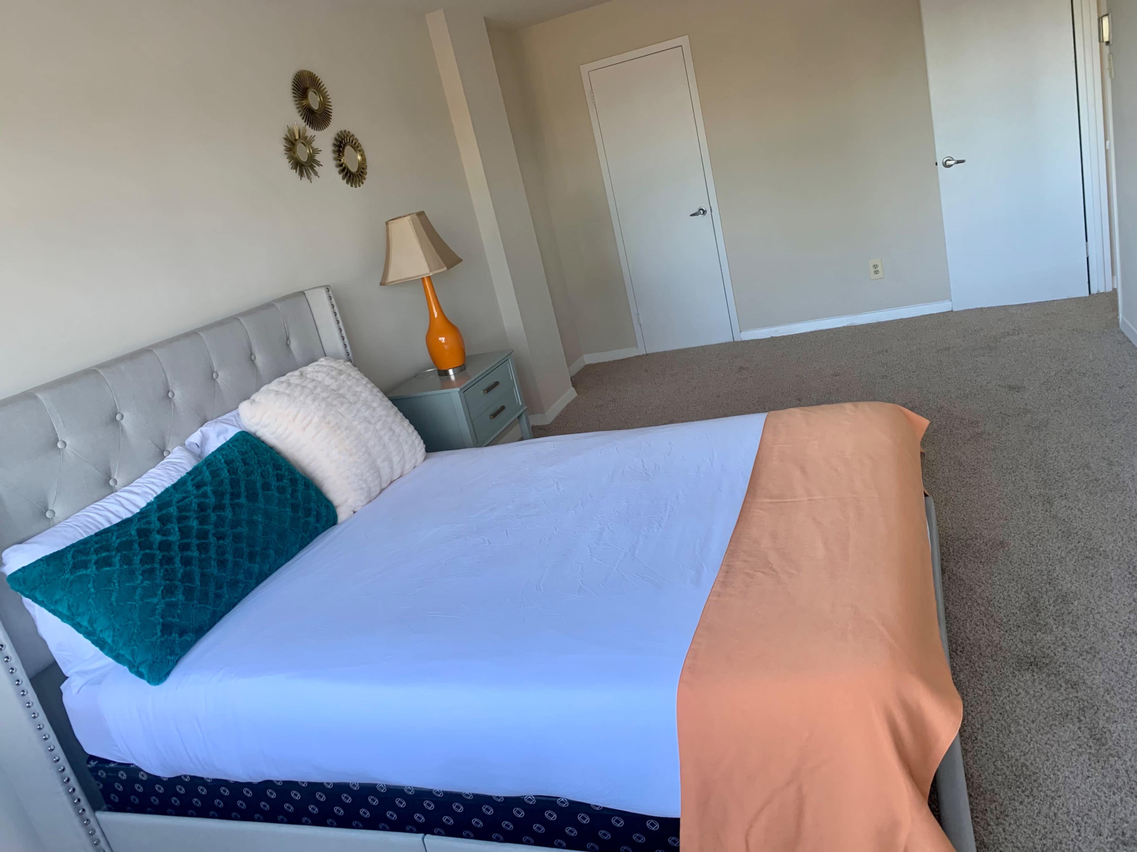 A neatly made bed with a blue and white decorative pillow, an orange throw blanket, a bedside lamp, and a dresser in a light-colored bedroom with carpeted flooring.