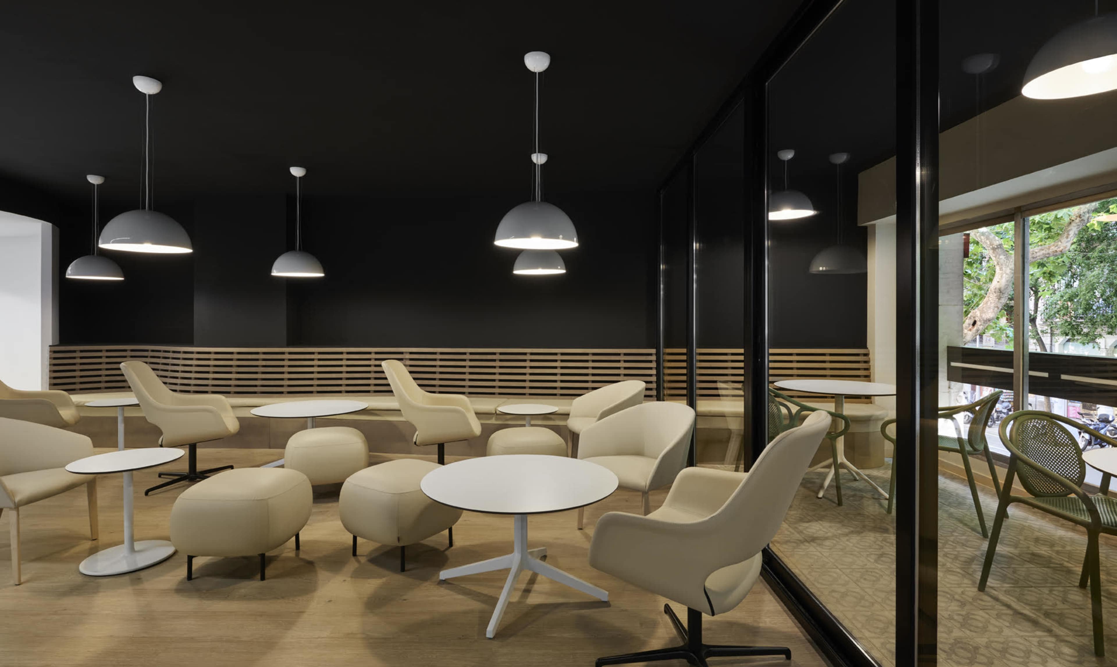 The image shows a modern lounge area featuring light-colored chairs and tables, with black walls and large windows allowing natural light to enter.