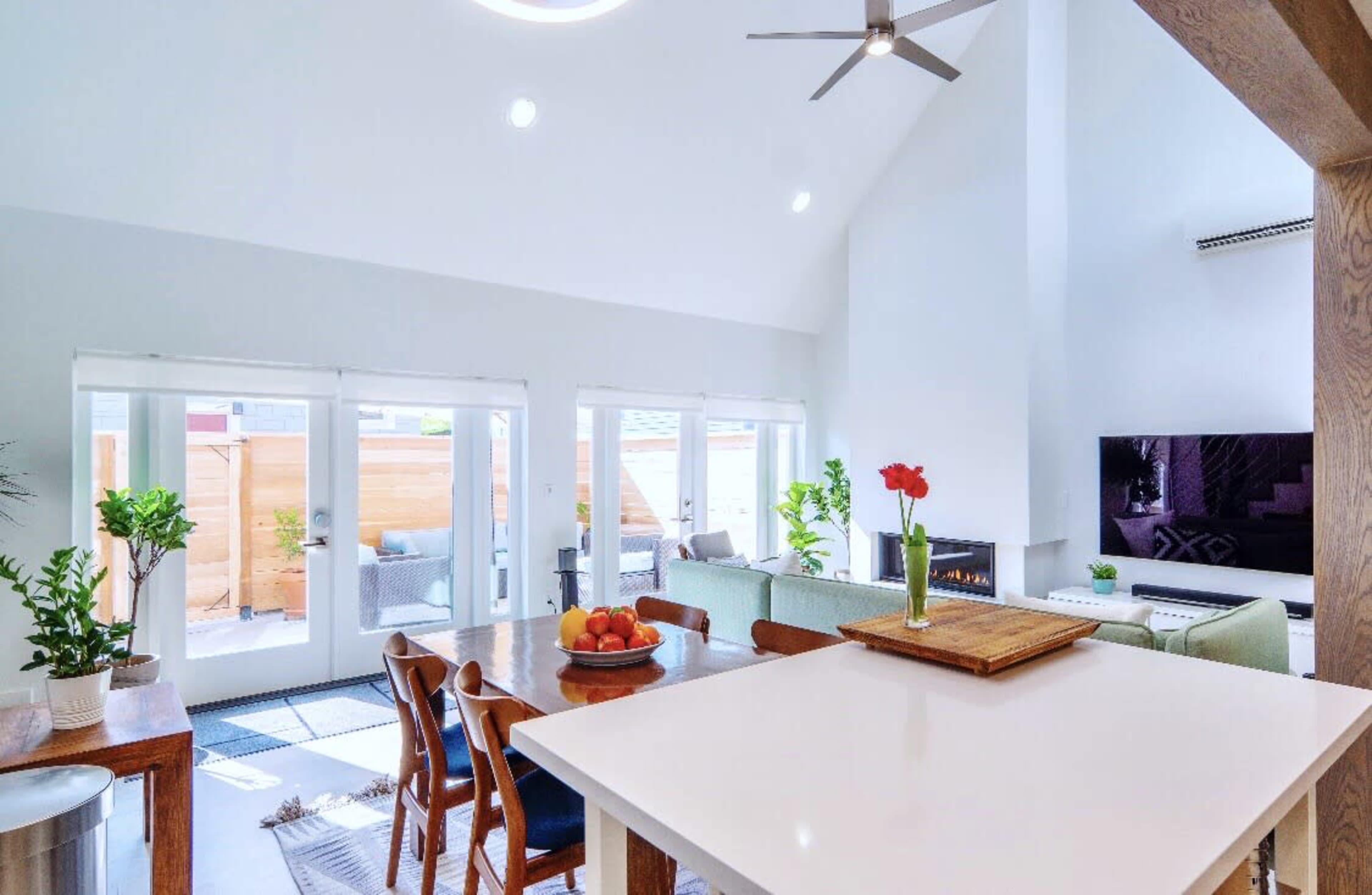 A modern open-concept living space features a dining area with a wooden table, a kitchen island, and large windows illuminating the room.