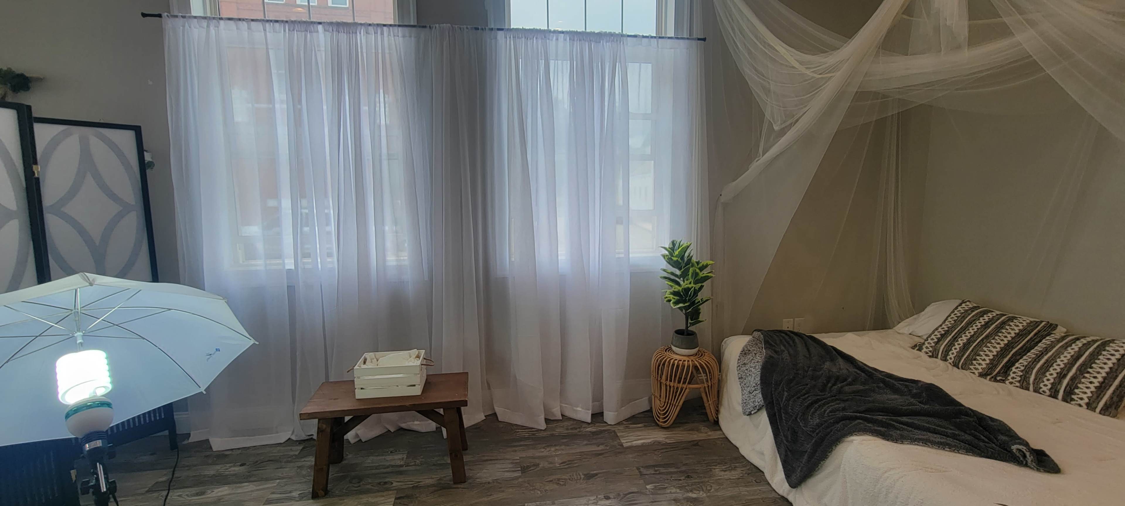 A well-lit room features a white bed with a blanket, a wooden table, a potted plant, and sheer curtains draping over a large window.