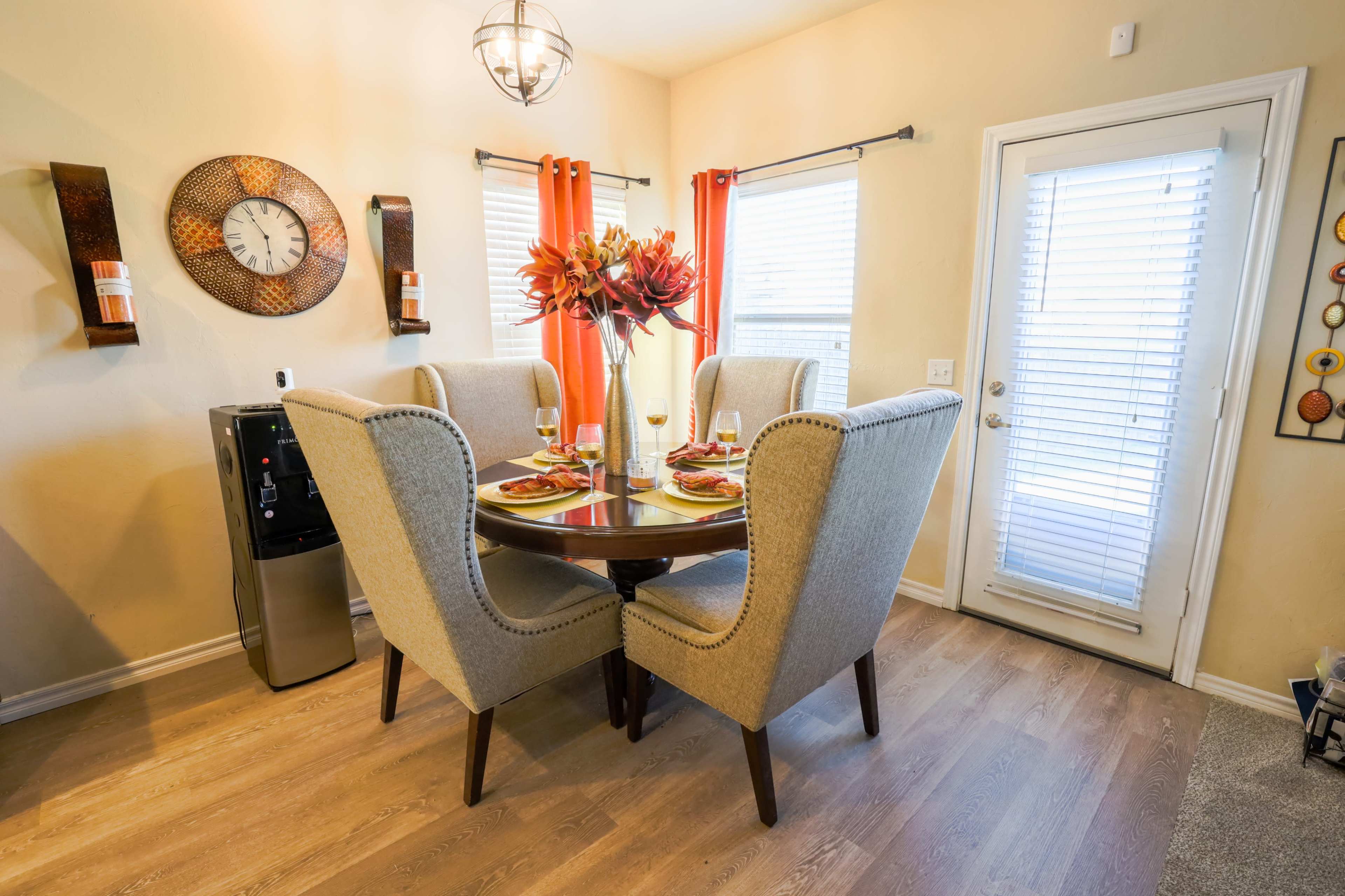 A dining area features a round table with two upholstered chairs, a vase of flowers, and a water dispenser against a wall with decorative accents.