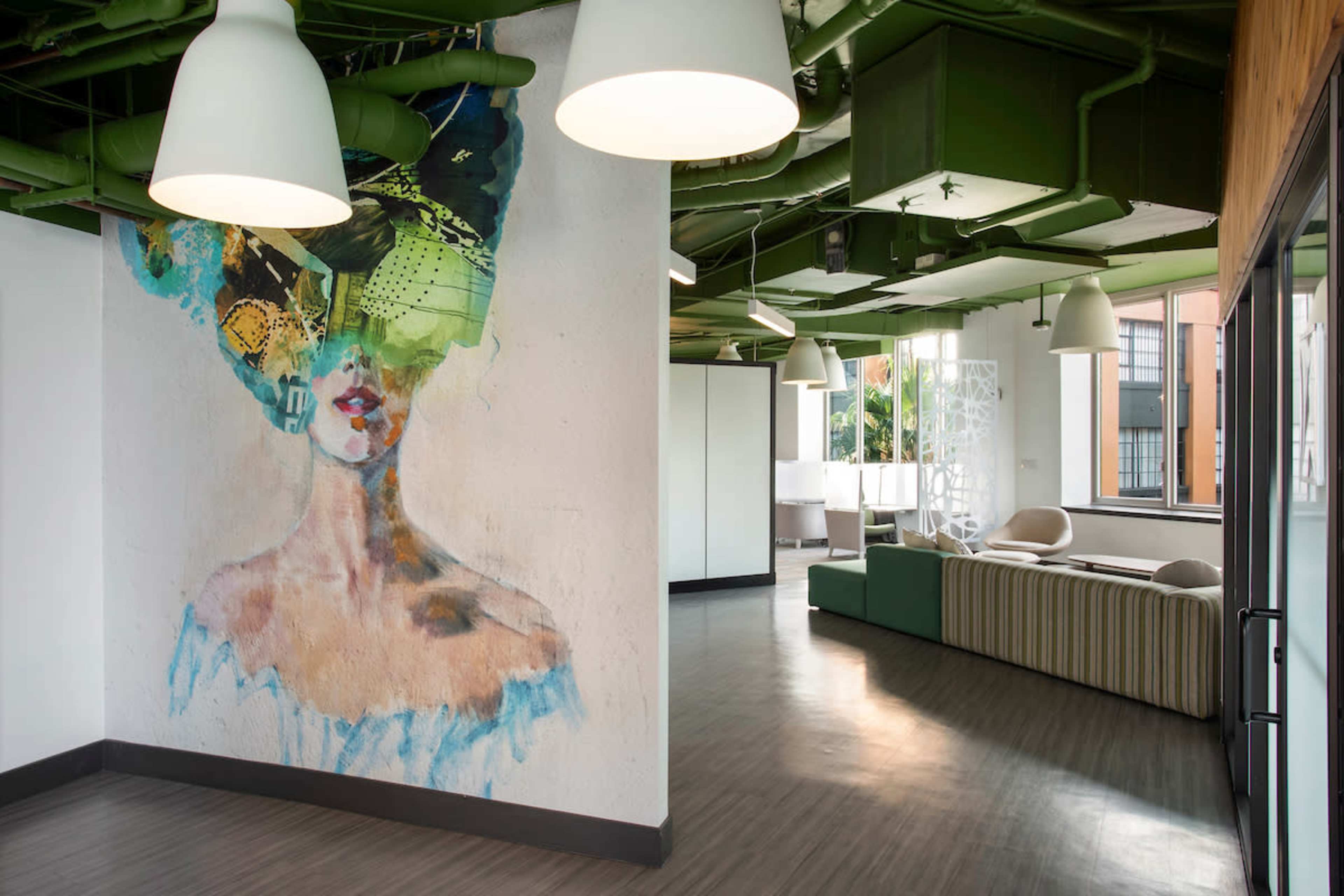 The image shows a modern interior space featuring a large mural of a woman's portrait on one wall, with green industrial-style ceiling pipes and contemporary furniture visible in the background.