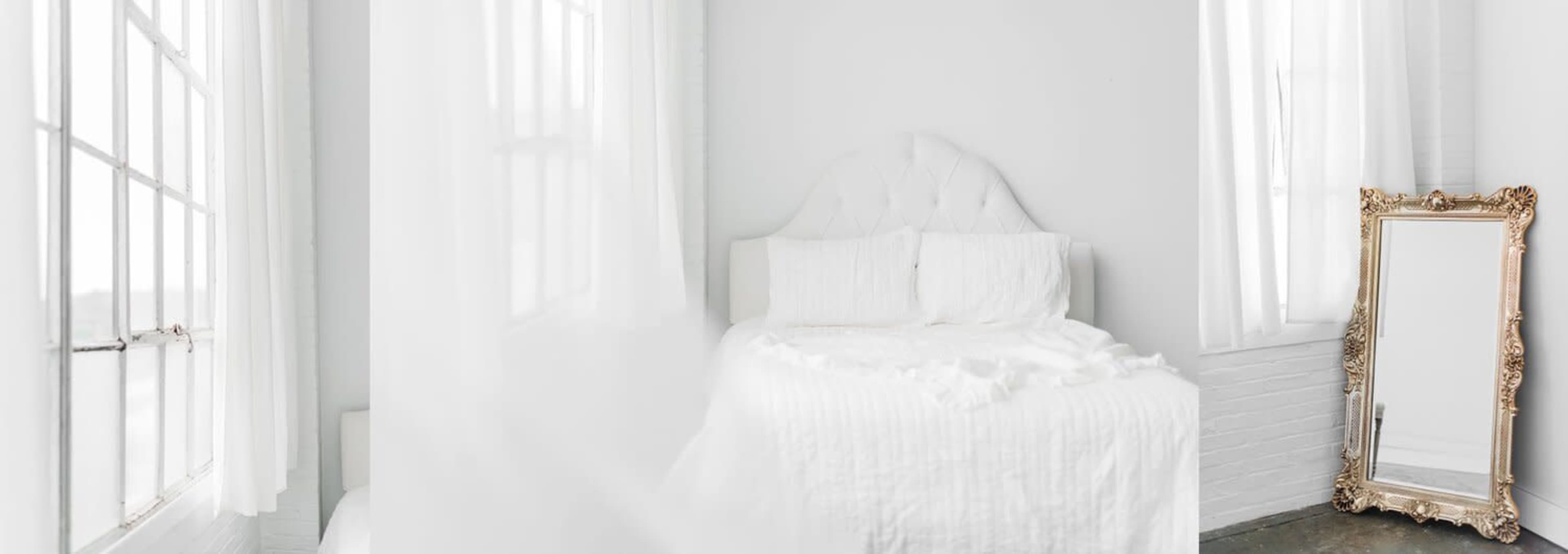 A bright bedroom features a white bed with light linens and a large, ornate mirror against the wall.
