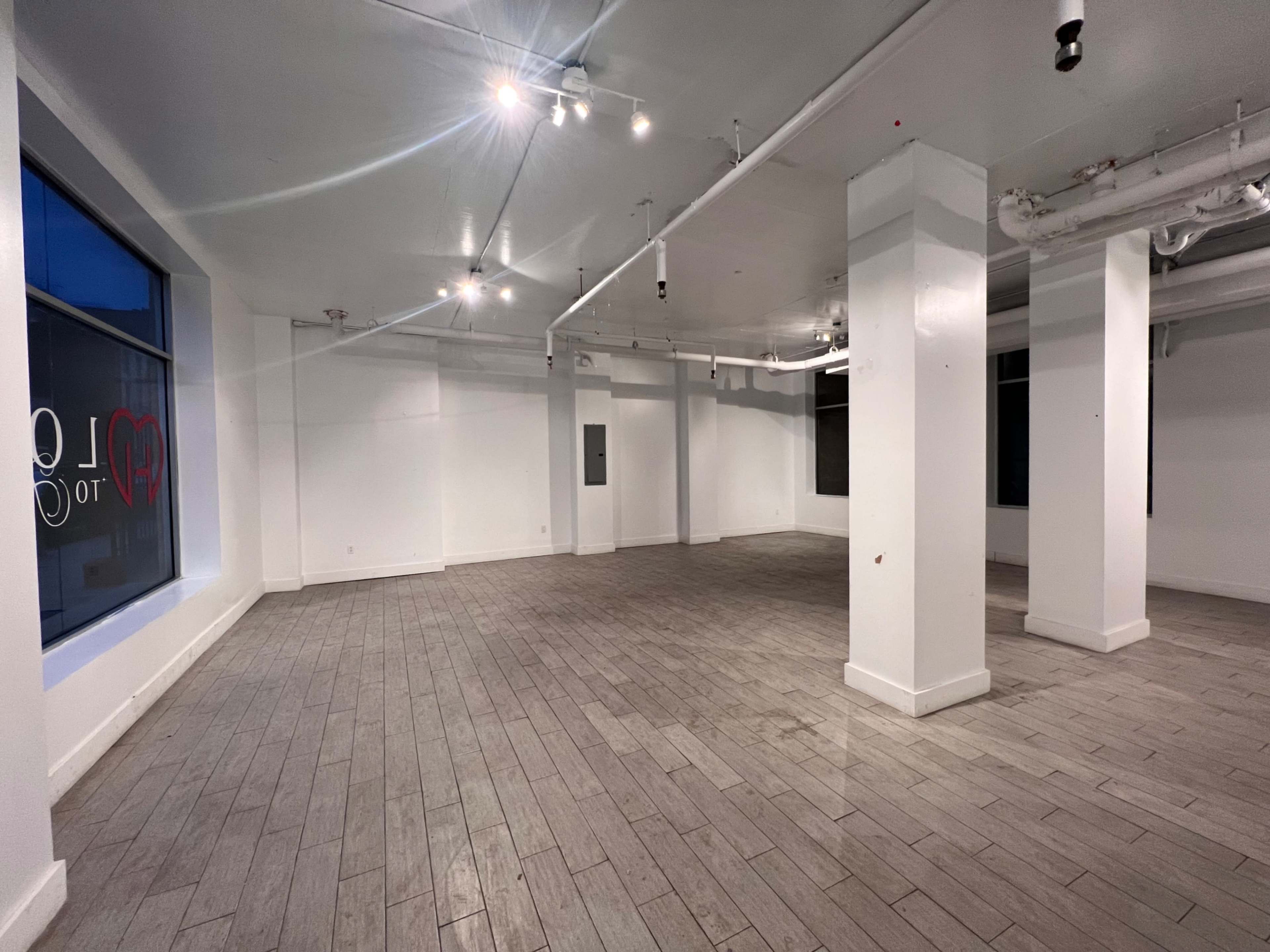 A spacious, empty room with white walls, gray wooden flooring, and several overhead lights.