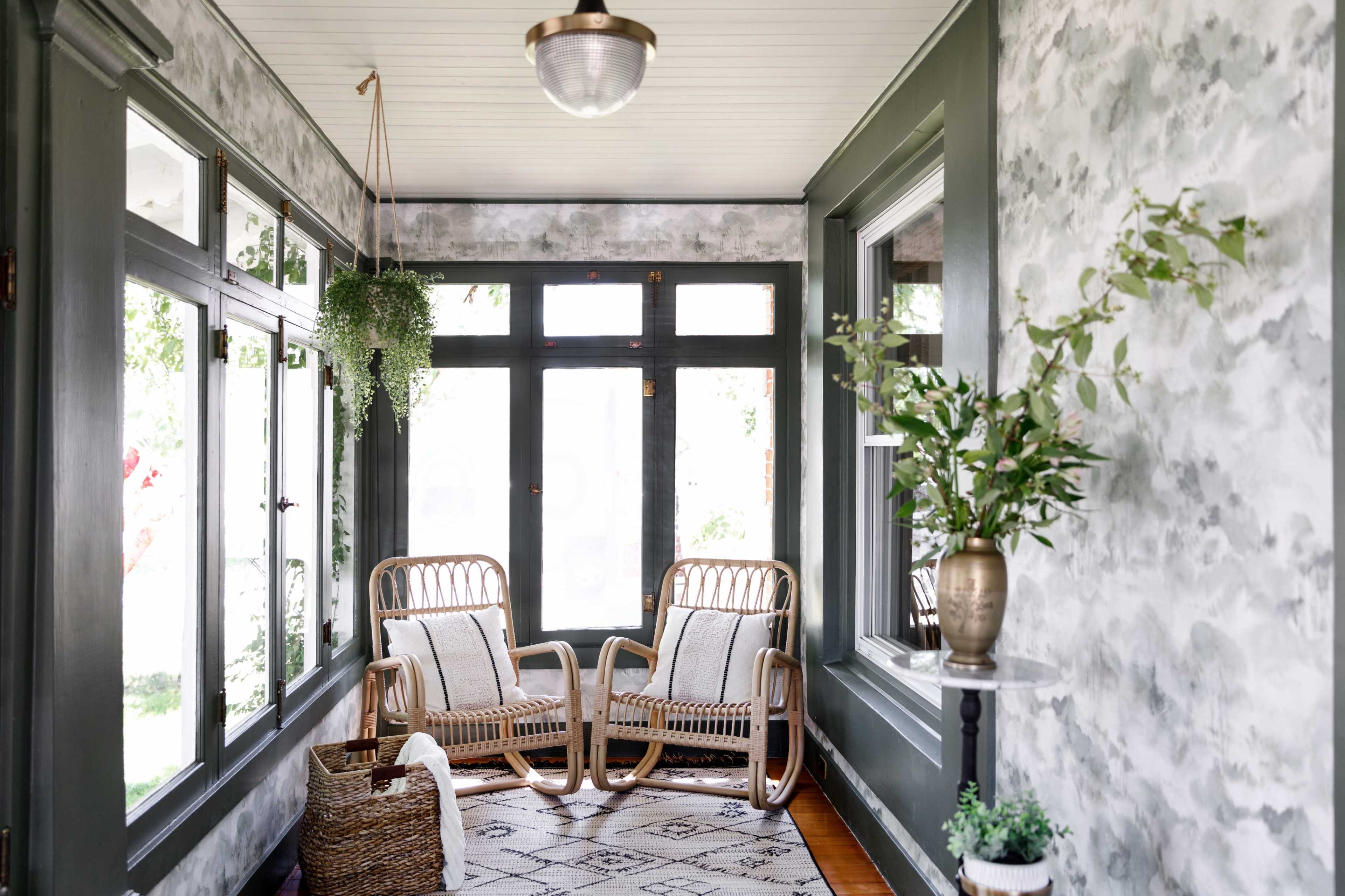 A sunlit room with large windows, featuring two wicker chairs, a potted plant, and a decorative vase atop a small table.
