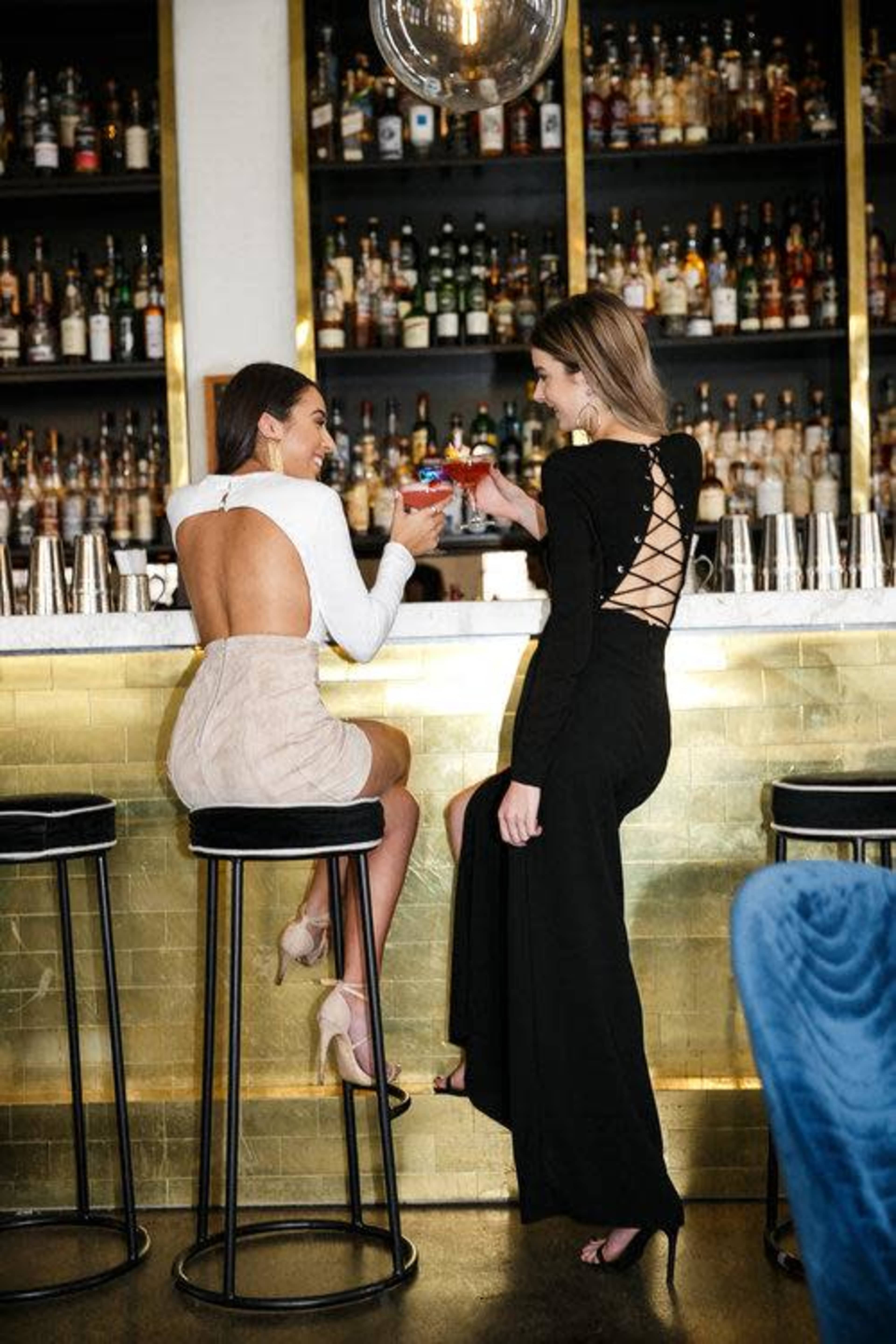 Two women in stylish evening dresses clink cocktail glasses while seated at a bar with a well-stocked shelf of liquor behind them.
