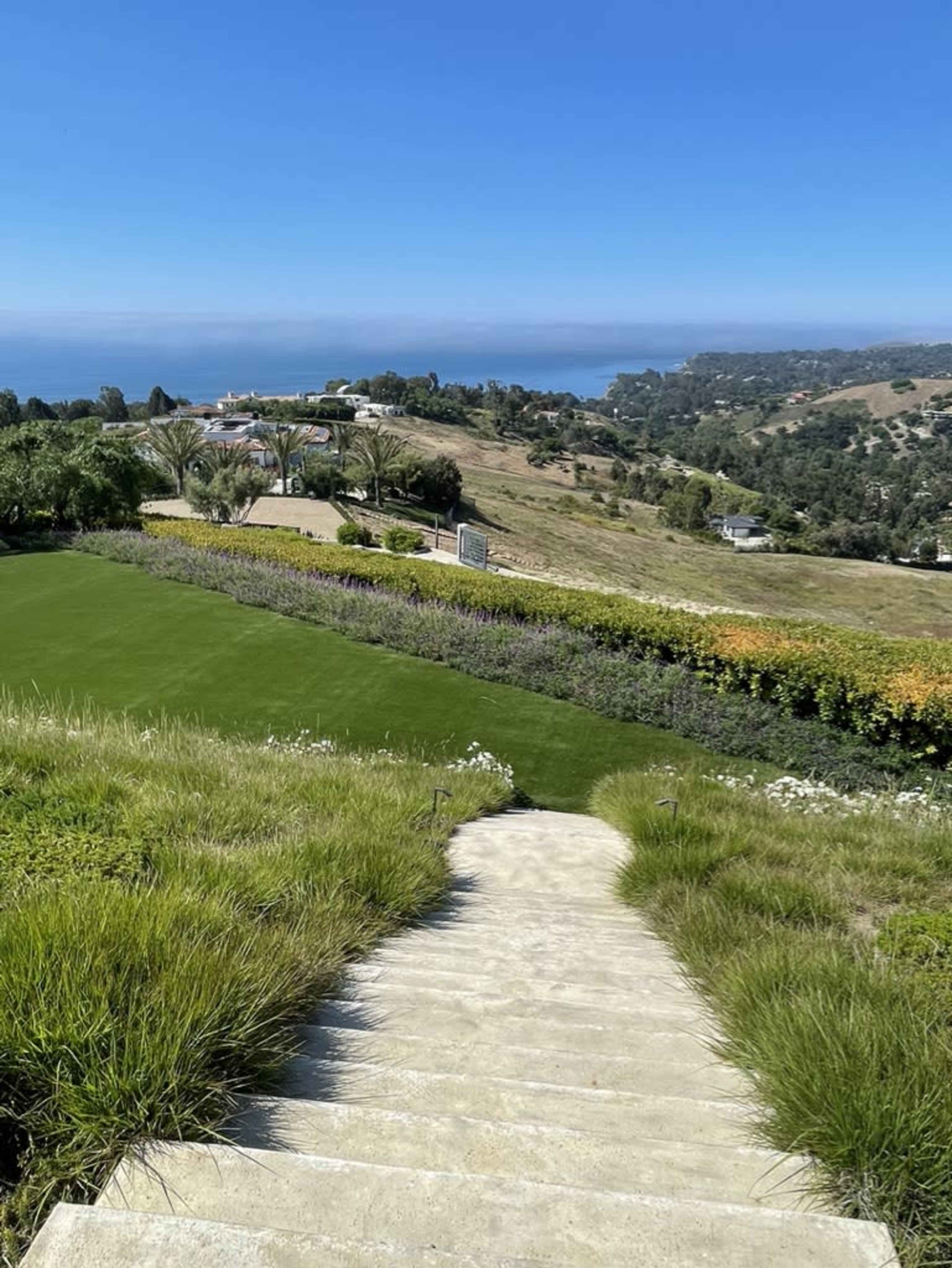 A stone staircase leading down to a lush green lawn, overlooking a scenic landscape with hills and the ocean in the distance.