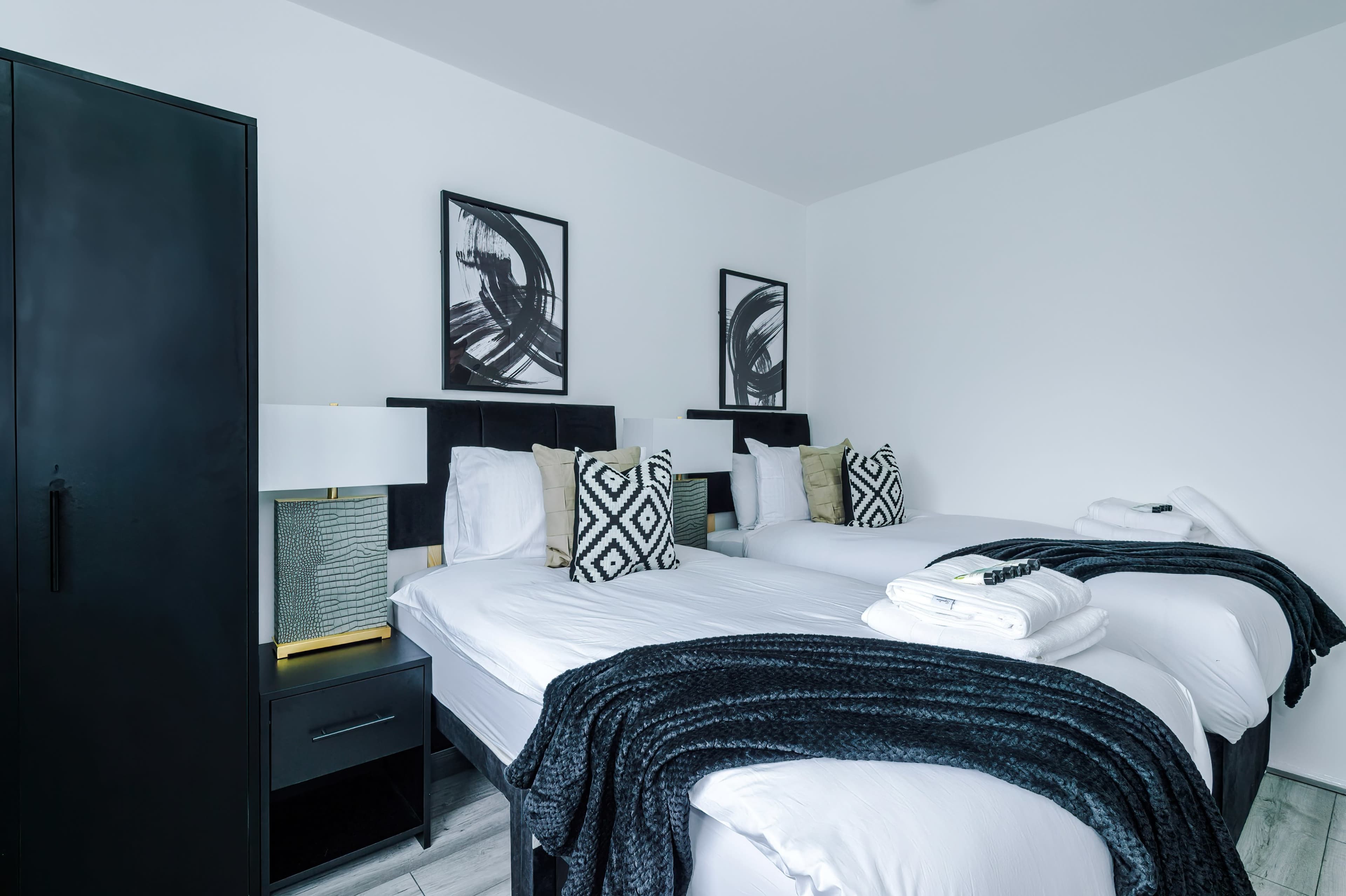 A modern hotel room featuring two twin beds, a black wardrobe, bedside tables with lamps, and framed artwork on the walls.