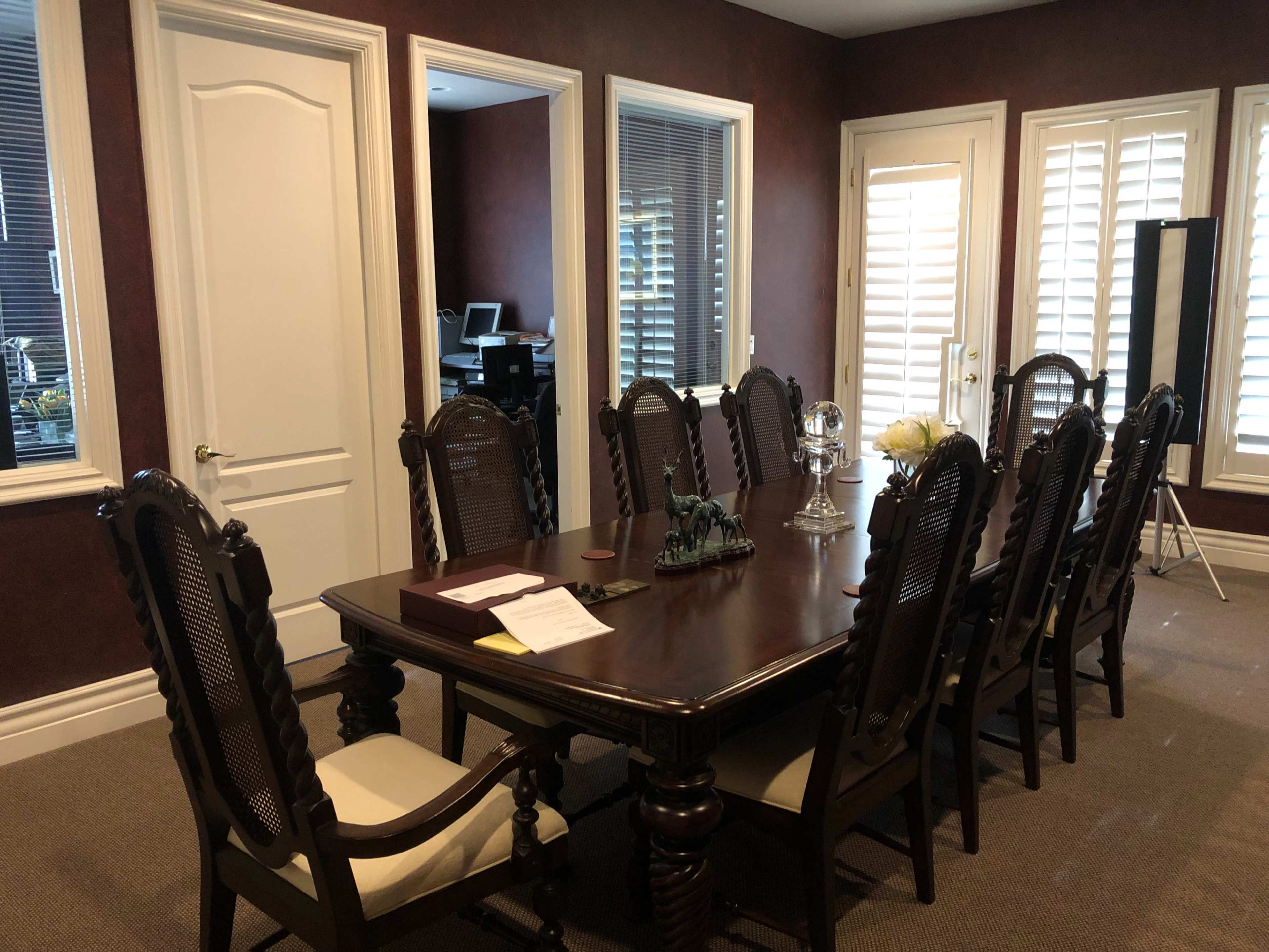 A large wooden conference table surrounded by eight chairs is positioned in a room with dark walls and several windows with blinds.