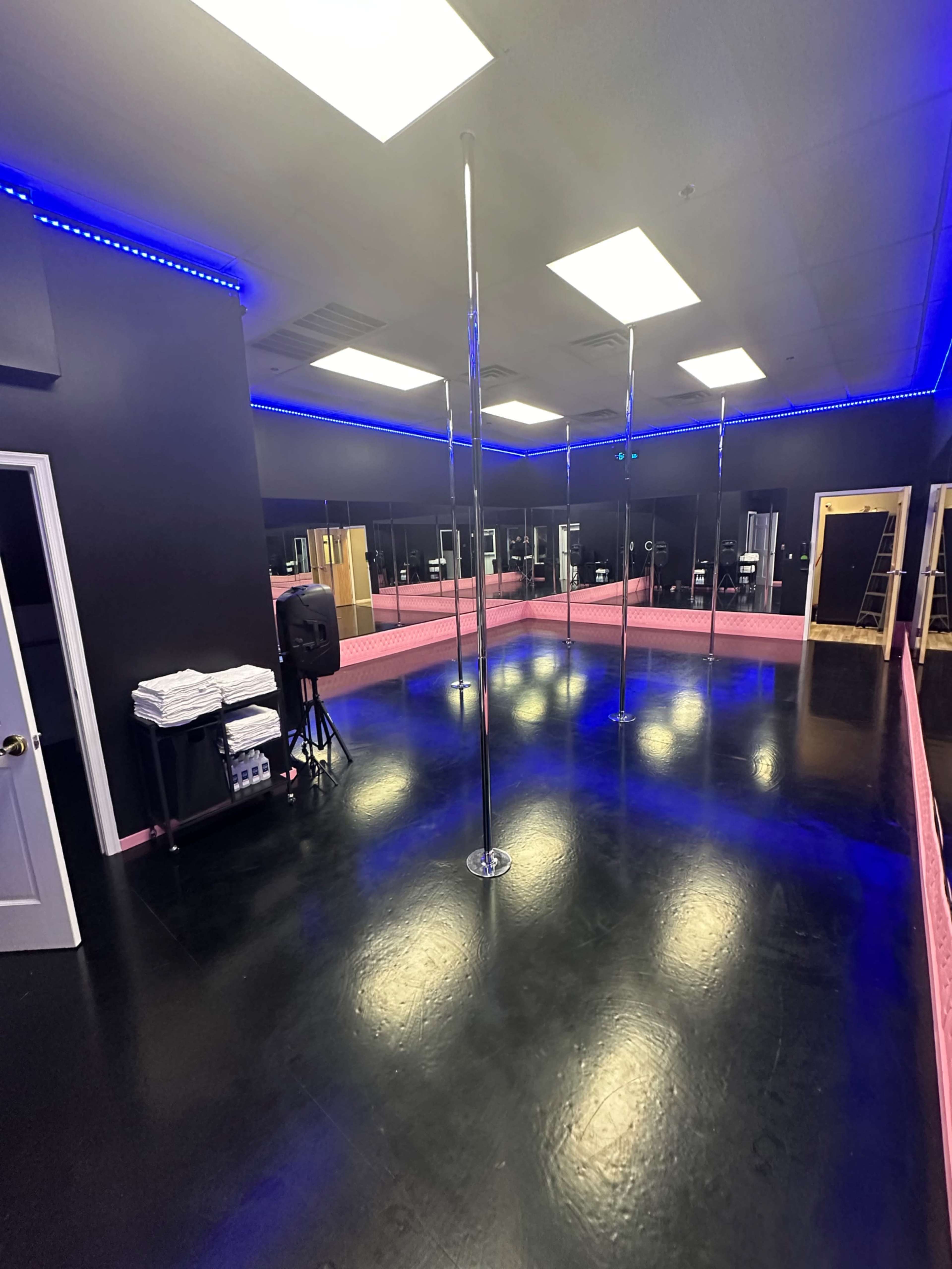 A spacious dance studio with a shiny black floor, mirrored walls, and several polished poles under blue LED lighting.
