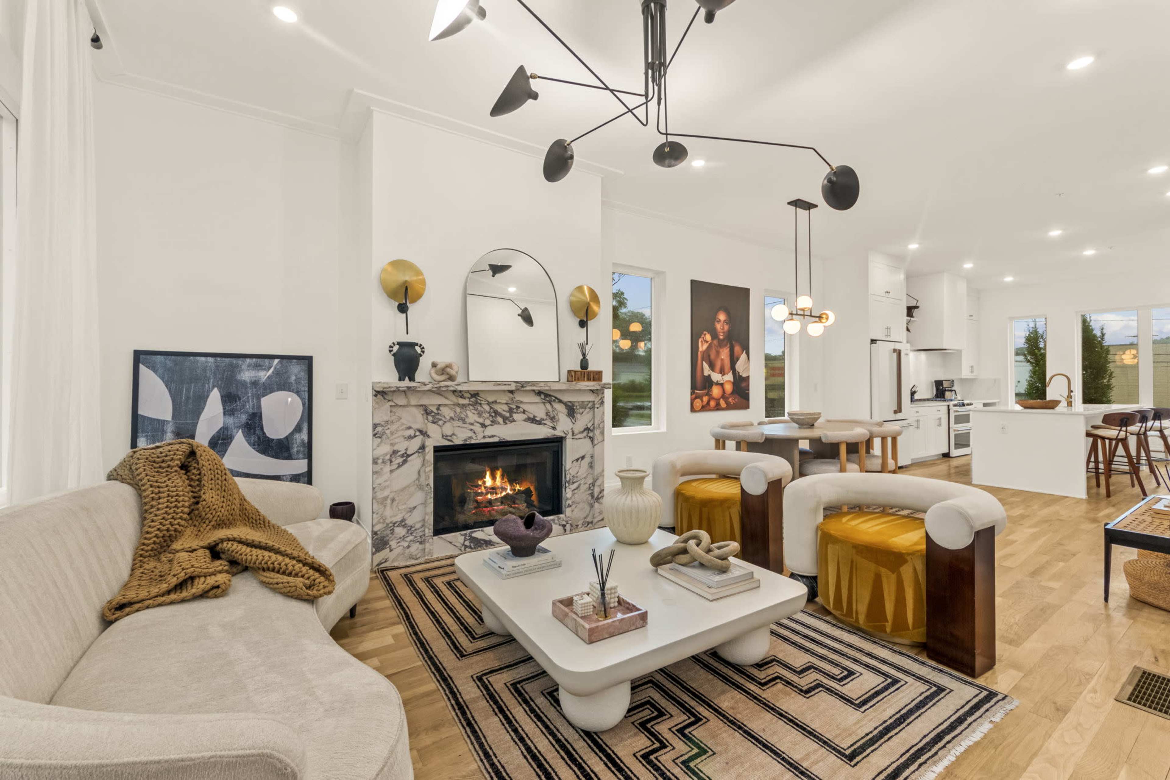 The image shows a modern living room with a marble fireplace, a circular mirror, a beige sofa, and a central coffee table surrounded by yellow accent chairs and a patterned rug.