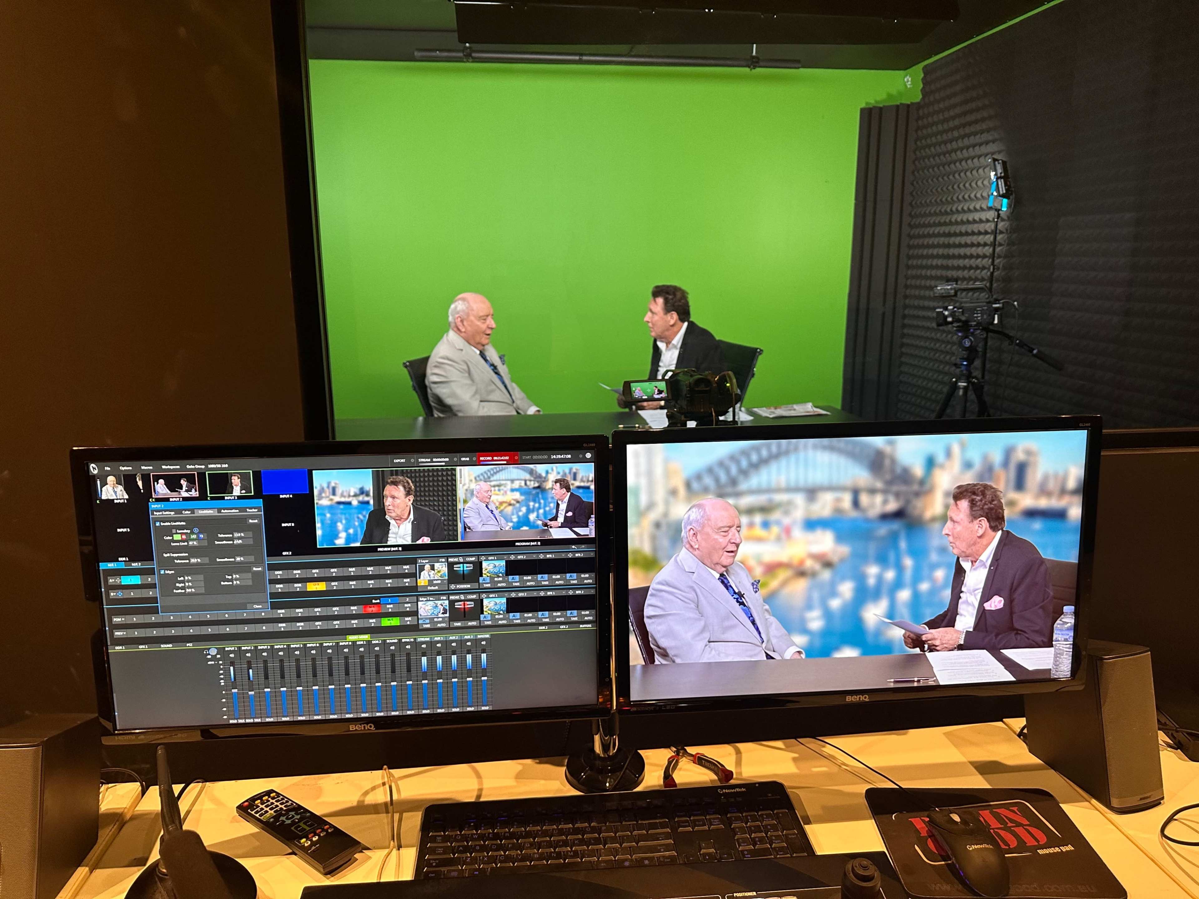 The image shows a video recording studio with two men sitting at a table, engaged in conversation, and two monitors displaying audio and video editing software.