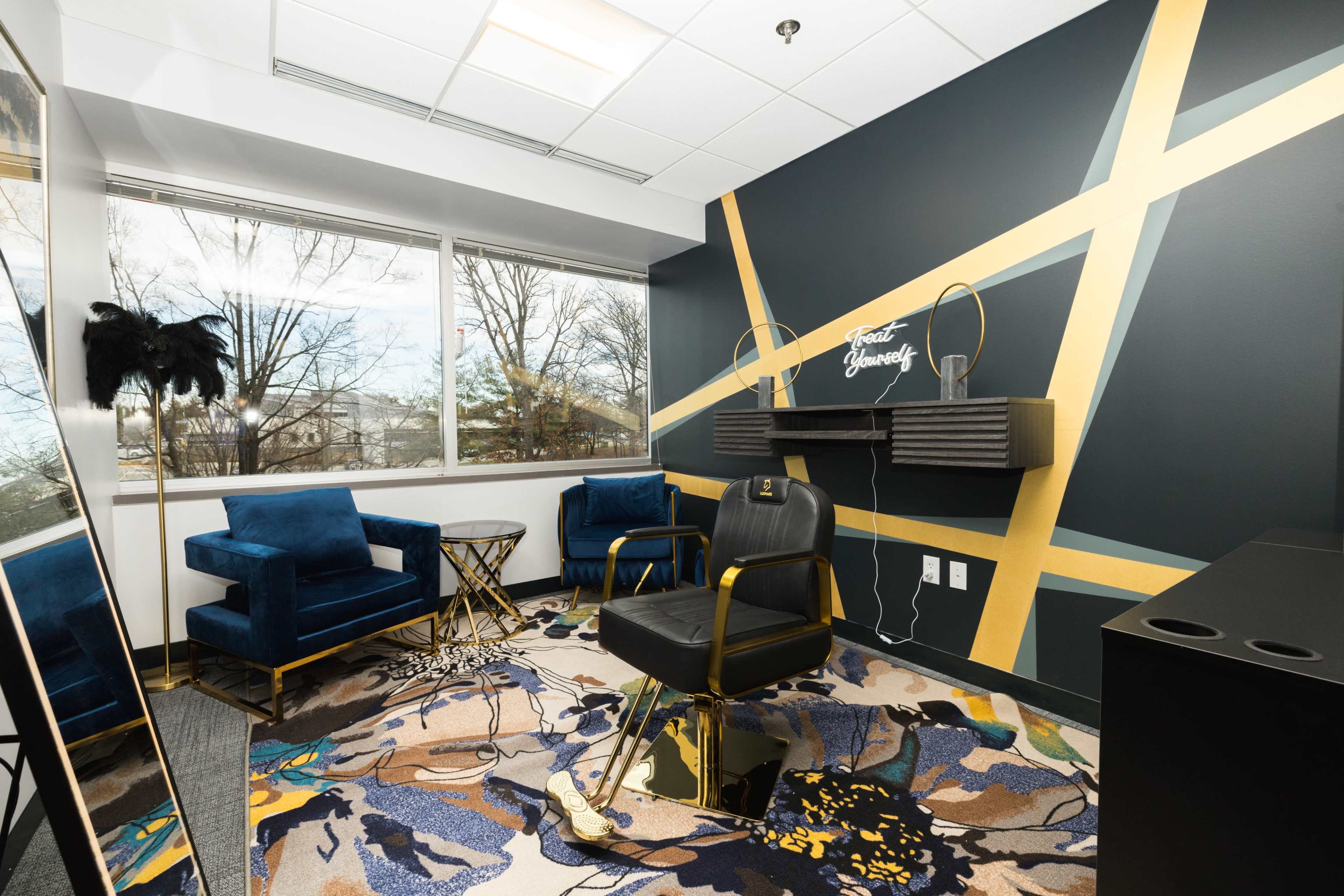 A modern lounge area with two blue velvet chairs, a round glass table, and a black office chair, set against a geometric gold-accented wall and a large window.