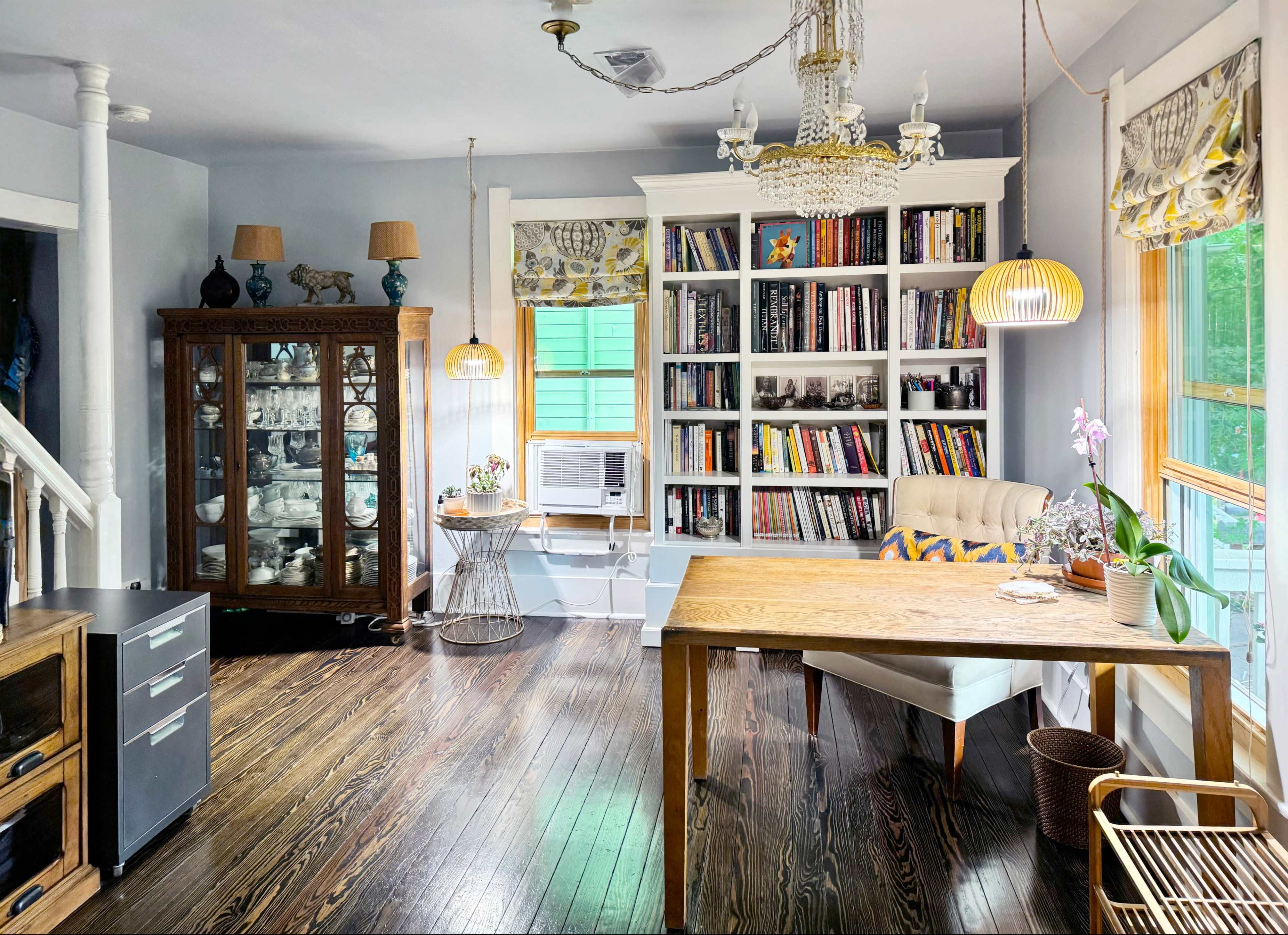 A cozy home office features a wooden desk, a bookshelf filled with books, a glass cabinet displaying ceramics, and an air conditioning unit near a window.