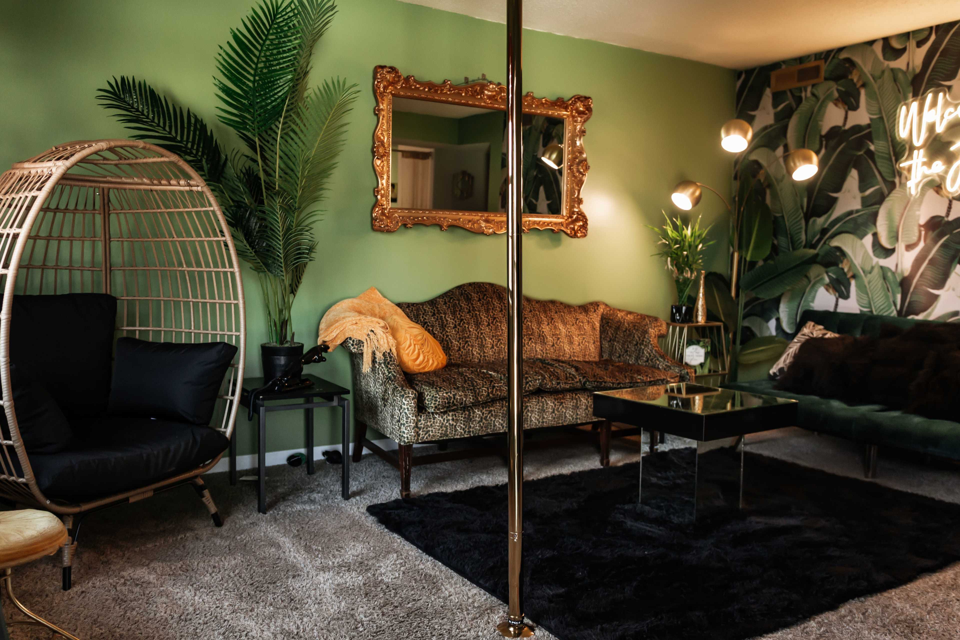 The image shows a stylish living room featuring a green wall, a retro-style sofa, a gold-framed mirror, and a tall plant alongside a modern black coffee table.