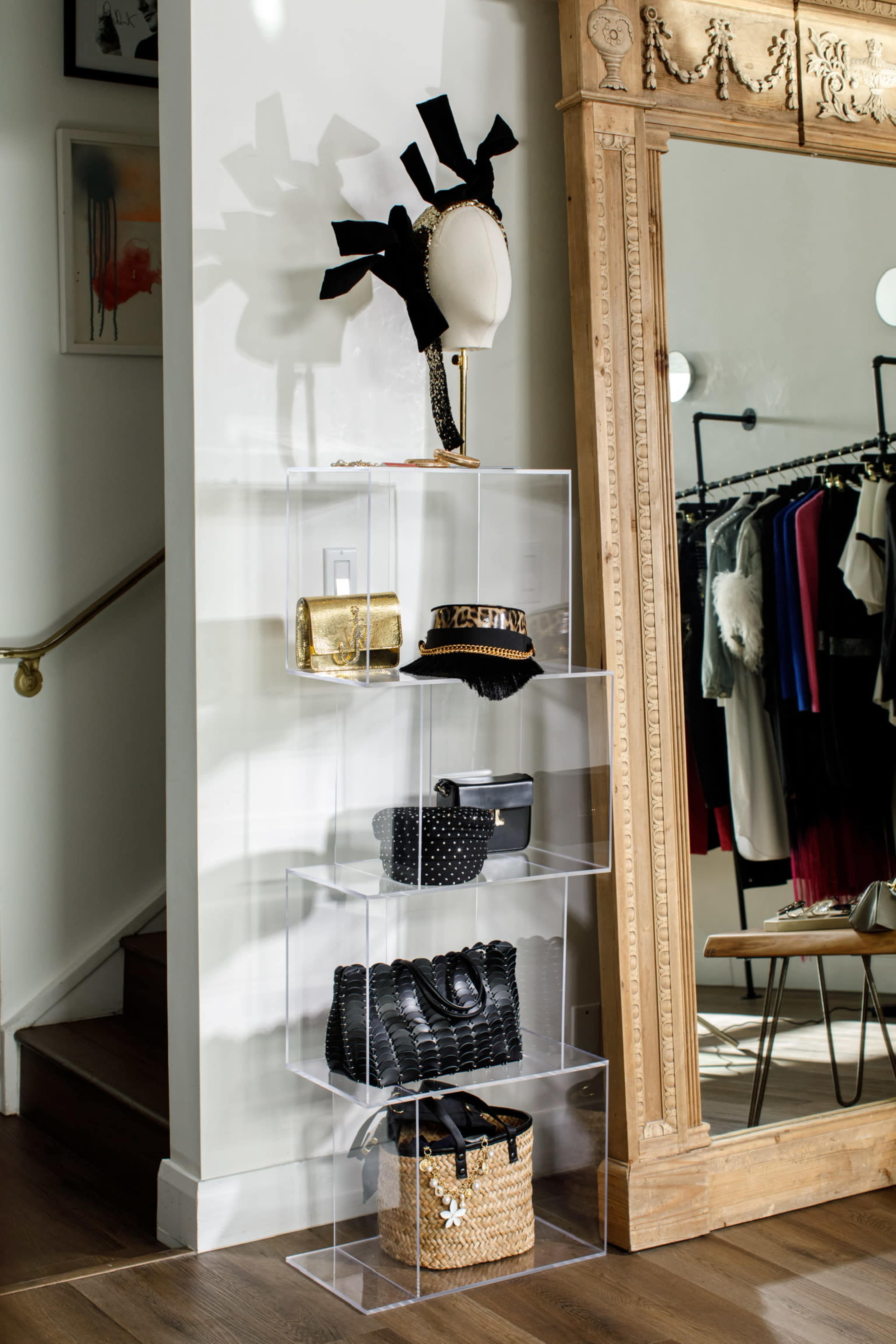 The image shows a clear display rack holding various handbags and a decorative hat next to a large mirror in a stylish boutique.