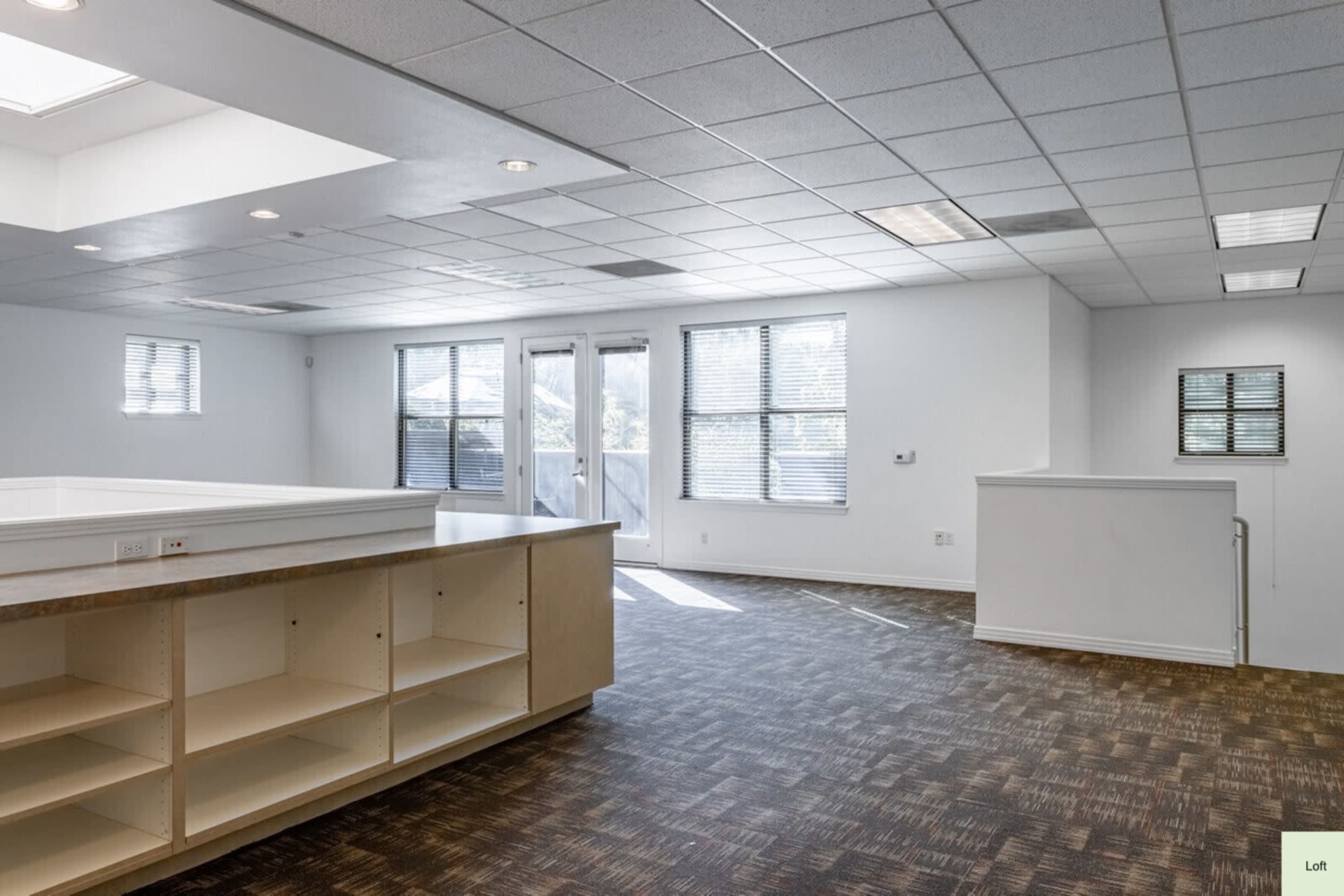 The image shows an empty room with large windows, a sliding glass door leading to a balcony, and a carpeted floor with an open area that includes built-in shelving.
