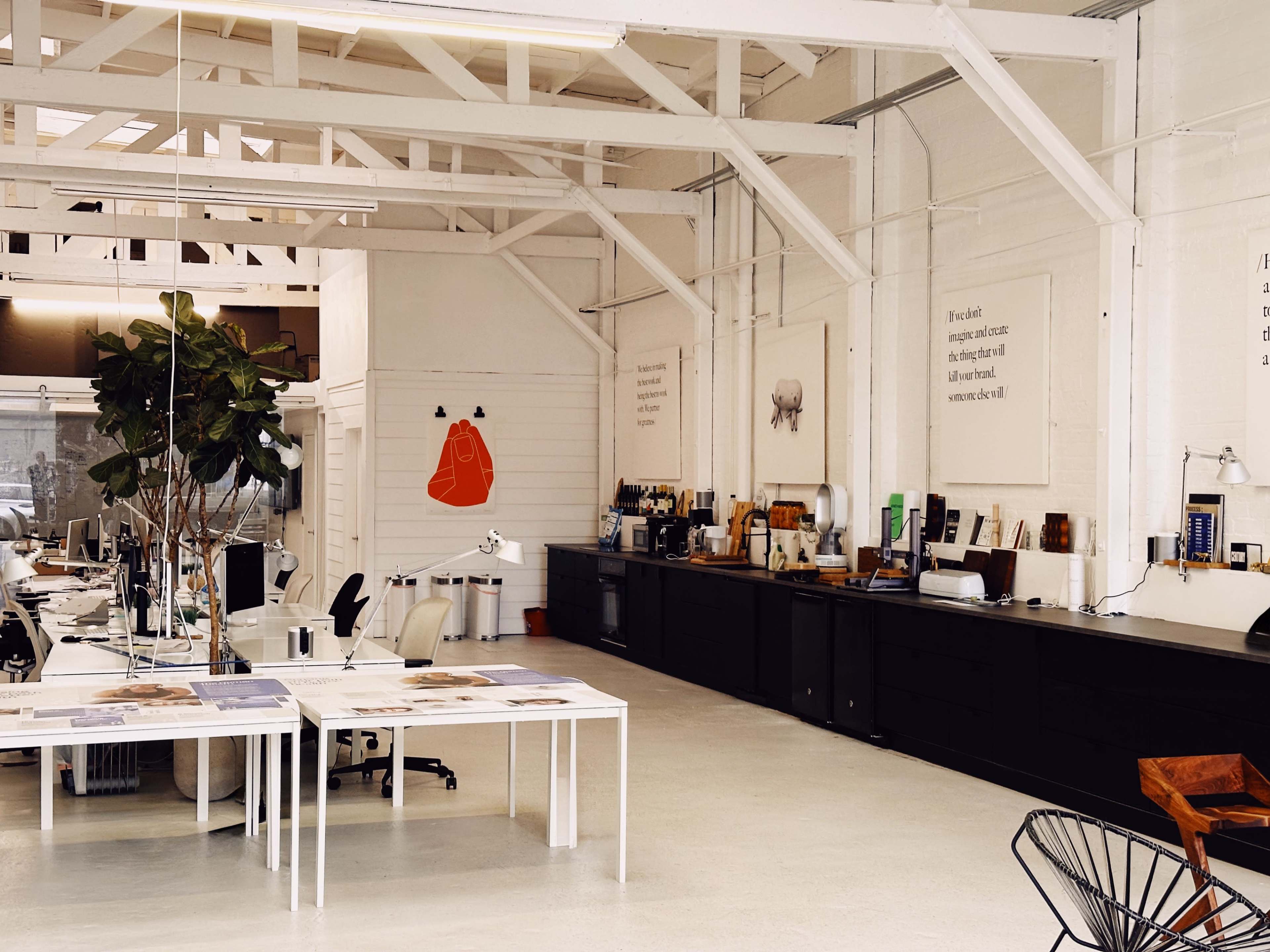 An open office space features white walls, large windows, wooden beams, and various workstations with minimalistic furniture.
