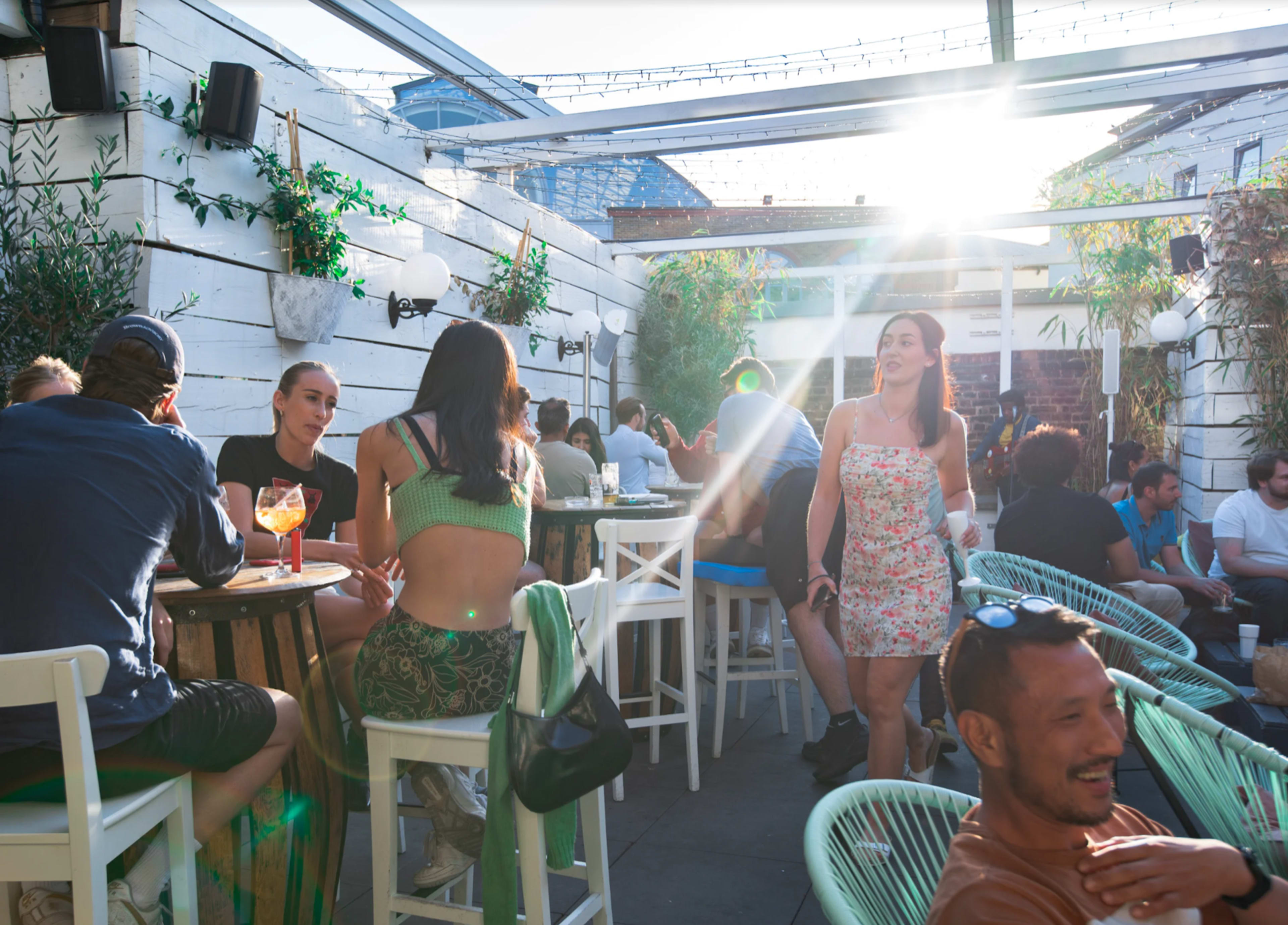 A lively outdoor bar scene features people dining and socializing under a glass roof with sunlight streaming through.