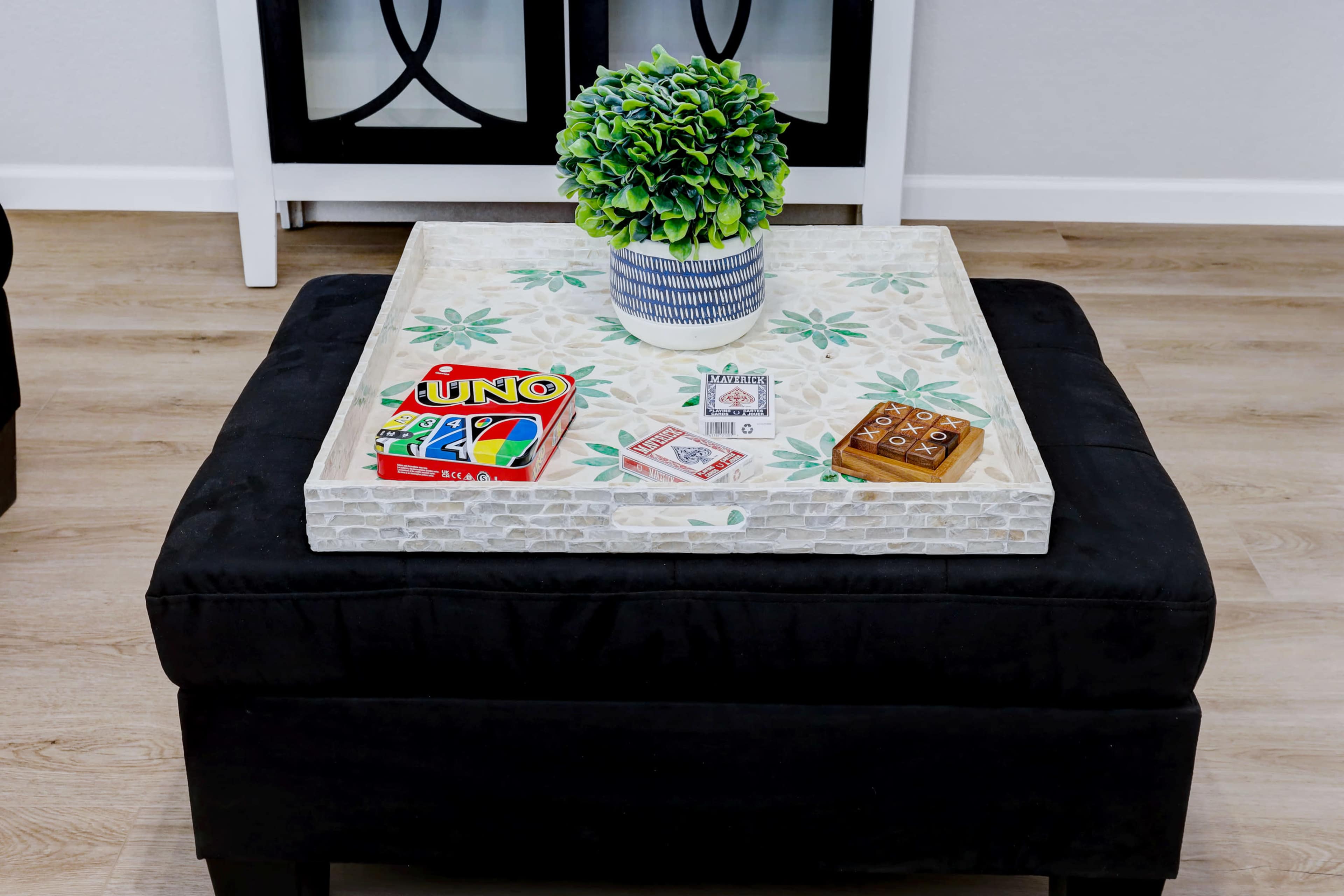 A square, patterned coffee table with a potted green plant and two card games resting on it sits in a modern living room space.