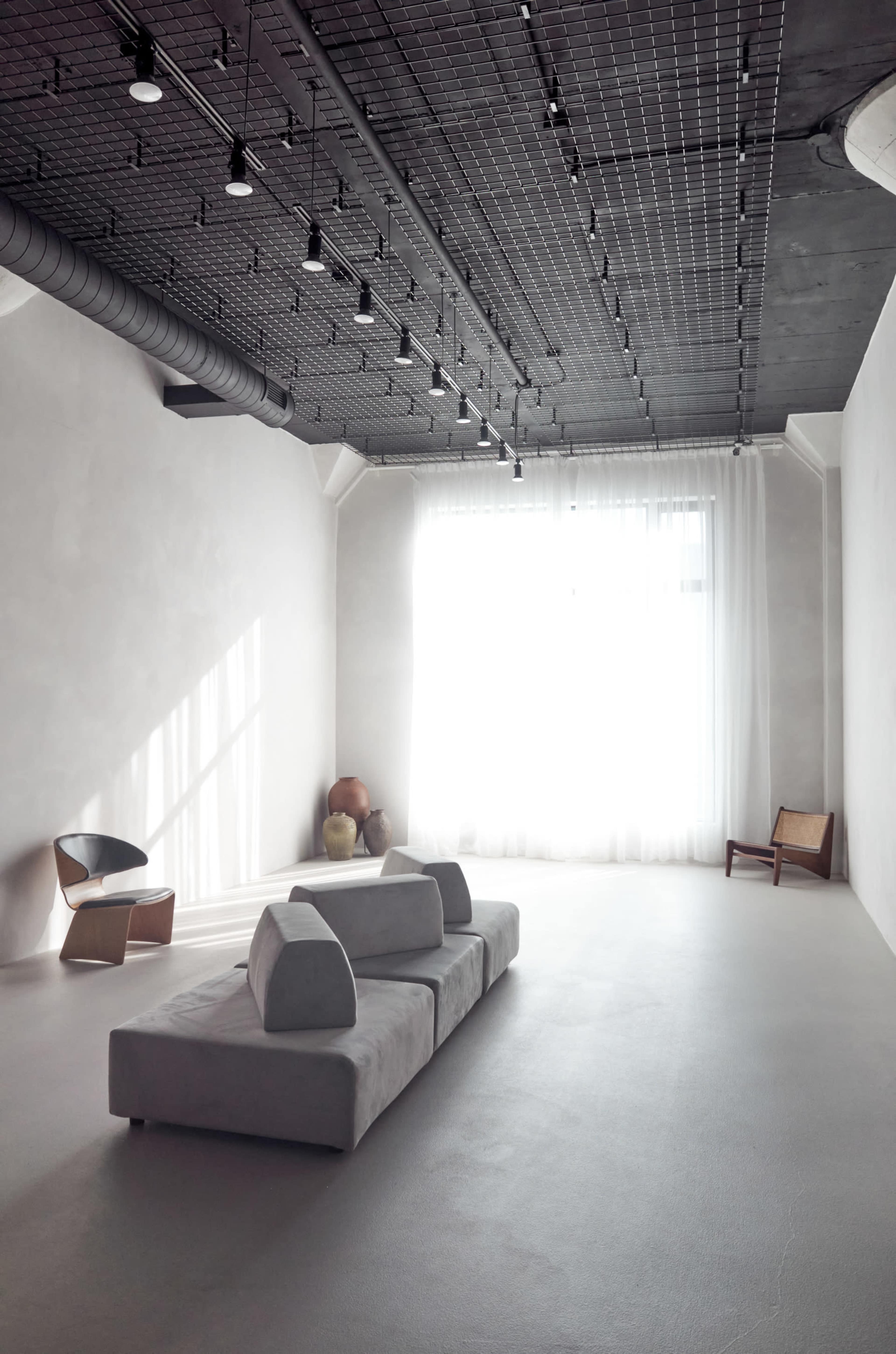 The image features a minimalist interior space with a light-colored floor, a low modern sofa in the center, and large windows covered by sheer curtains allowing natural light to enter.