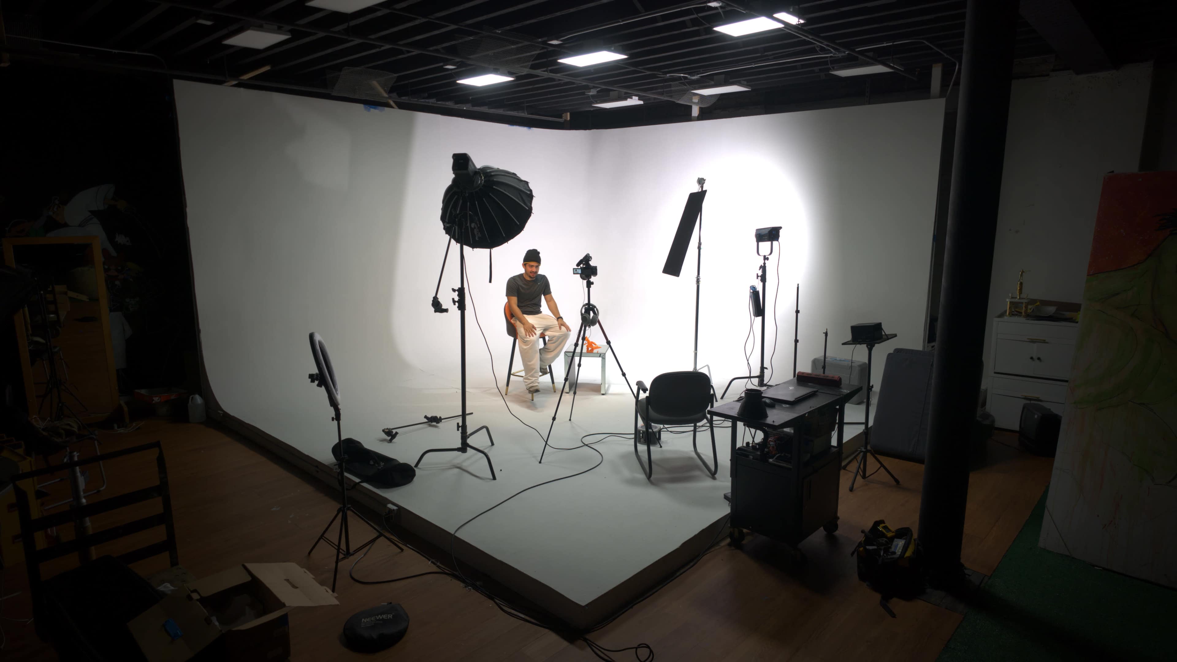 A person sits in a chair in a well-lit studio surrounded by various lighting equipment and camera gear.