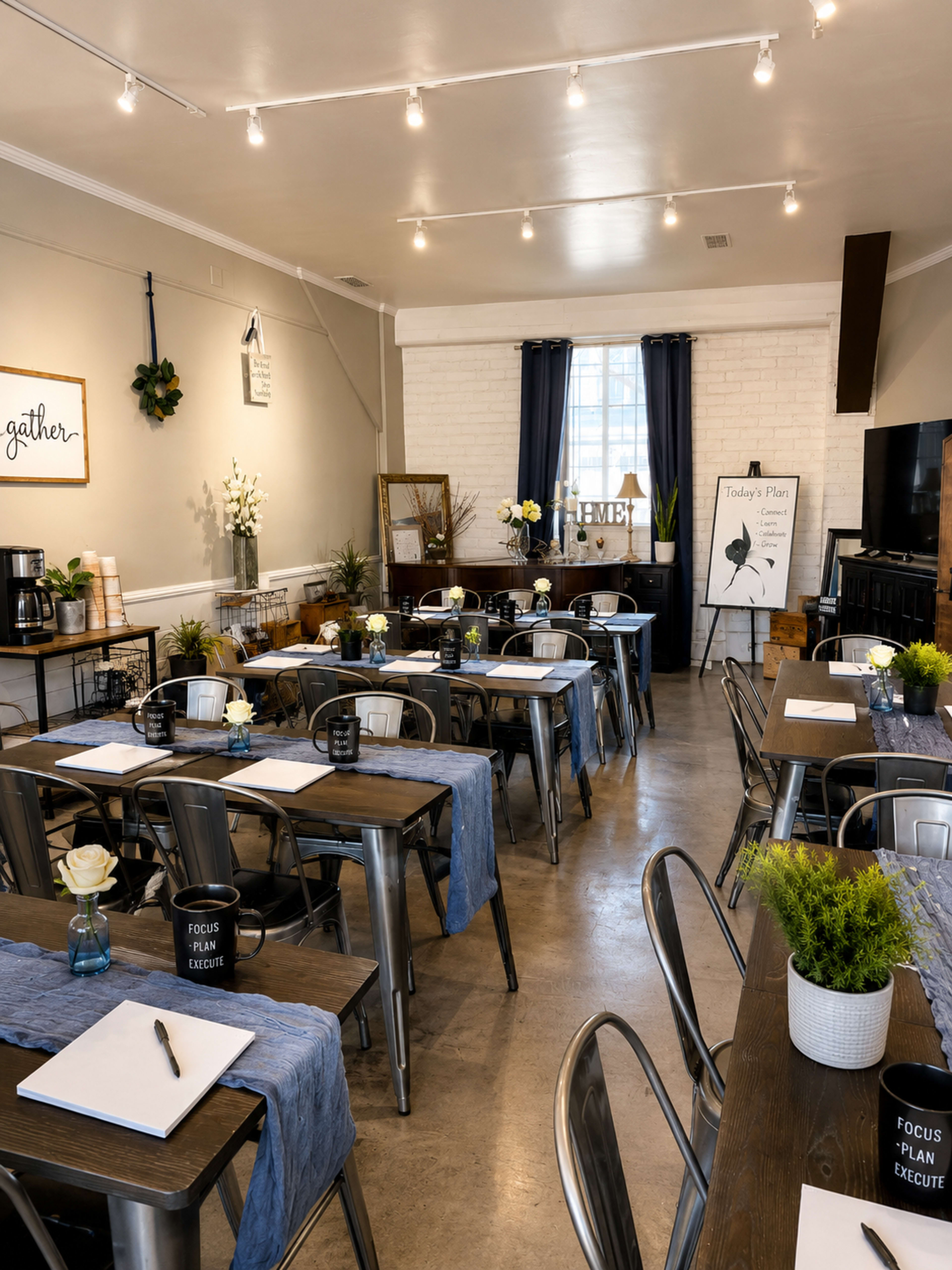 The image shows a neatly arranged meeting room with several tables set for a presentation, featuring blue table runners, floral centerpieces, and a large chalkboard displaying an agenda.