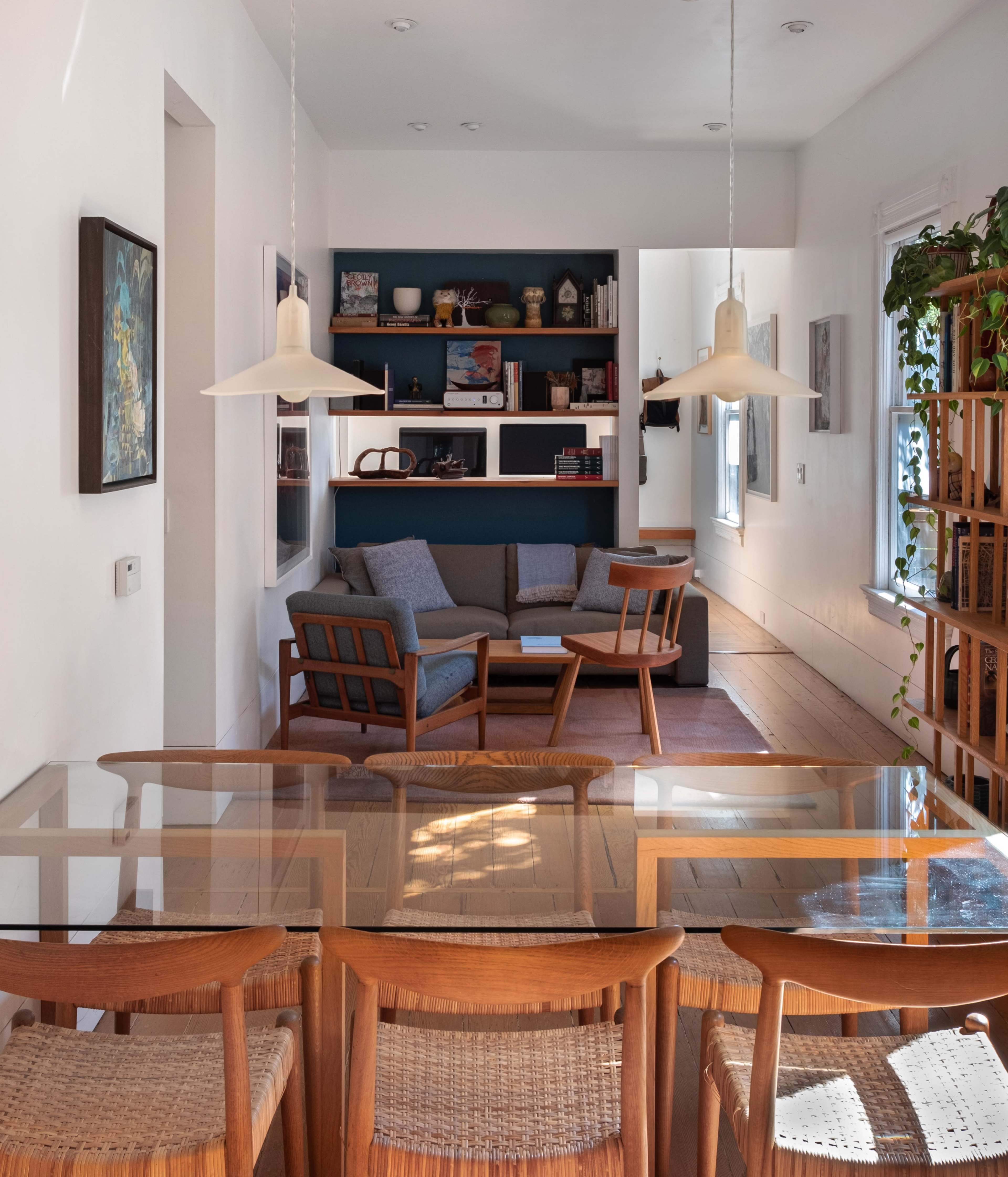 A dining area with a glass-top table and wooden chairs overlooks a living space featuring a sofa, bookshelf, and large windows.