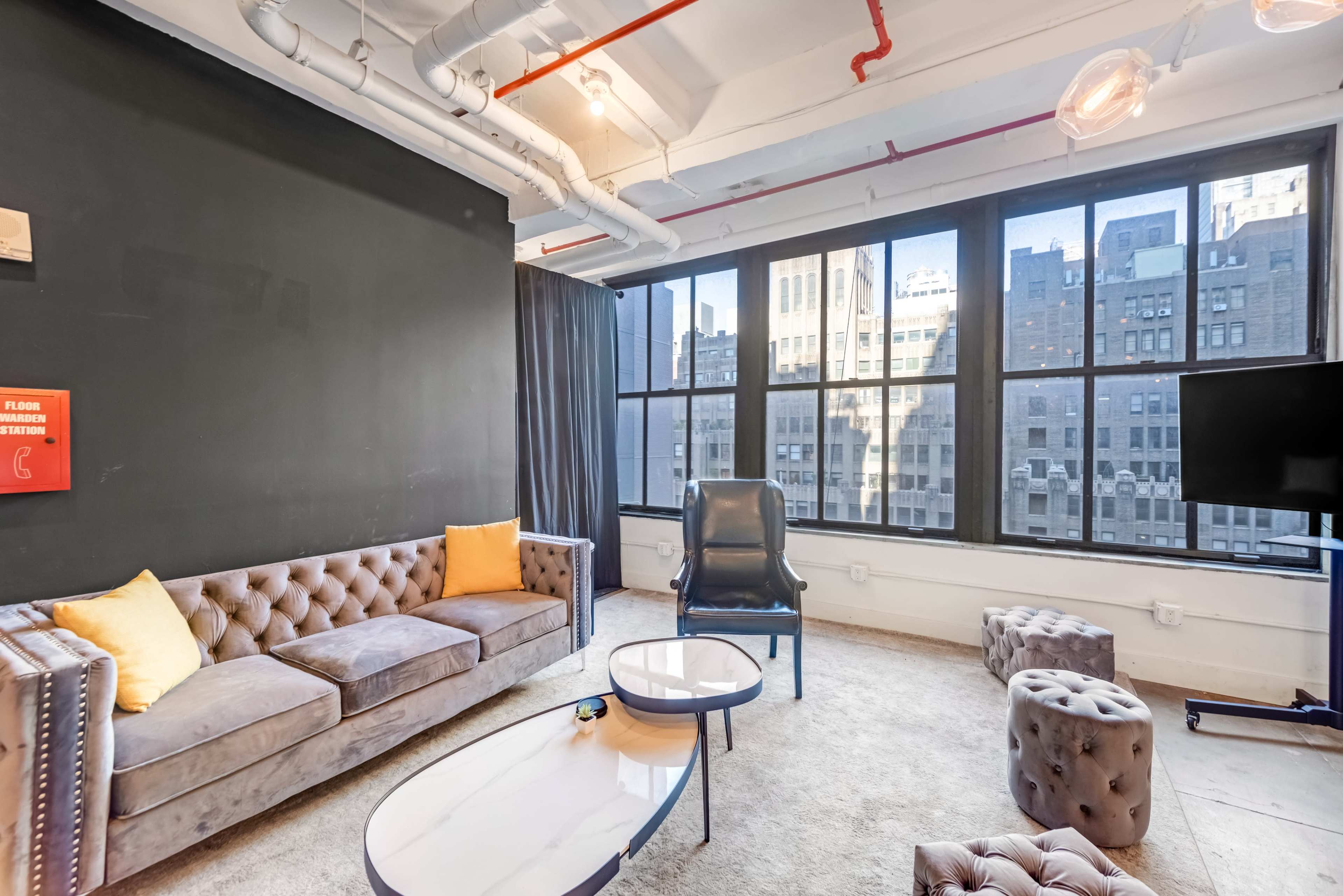 A modern lounge area with a tufted gray sofa, two ottomans, and large windows overlooking a cityscape.