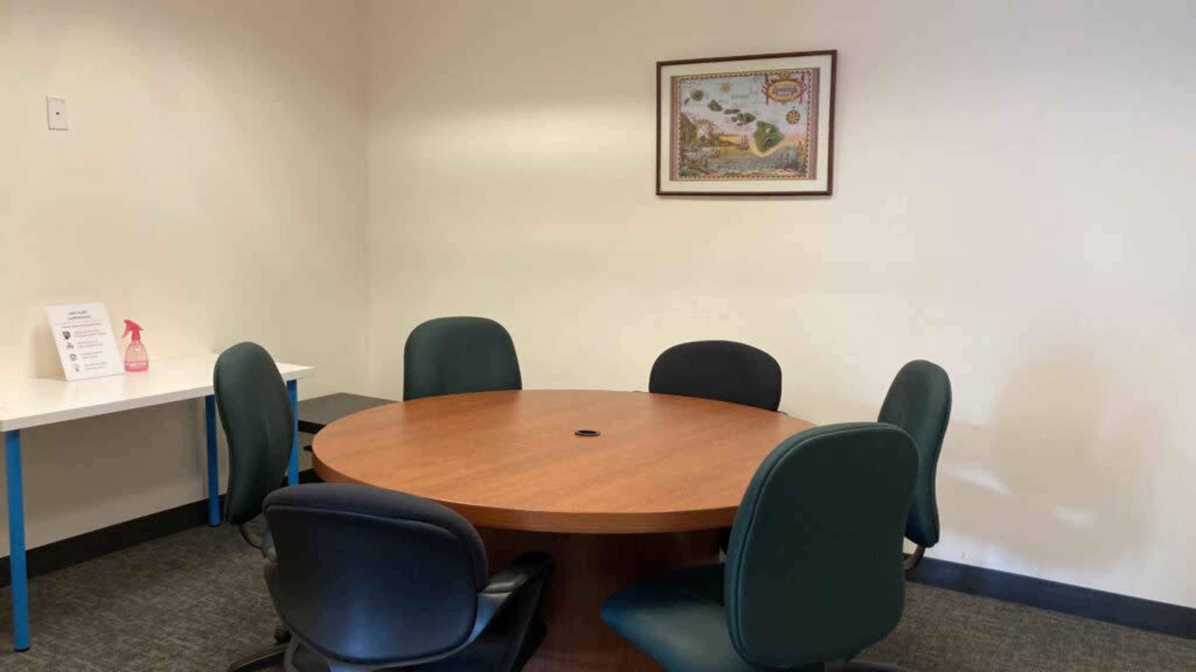 A round wooden conference table is surrounded by five green chairs in a plain meeting room with a framed picture on the wall.