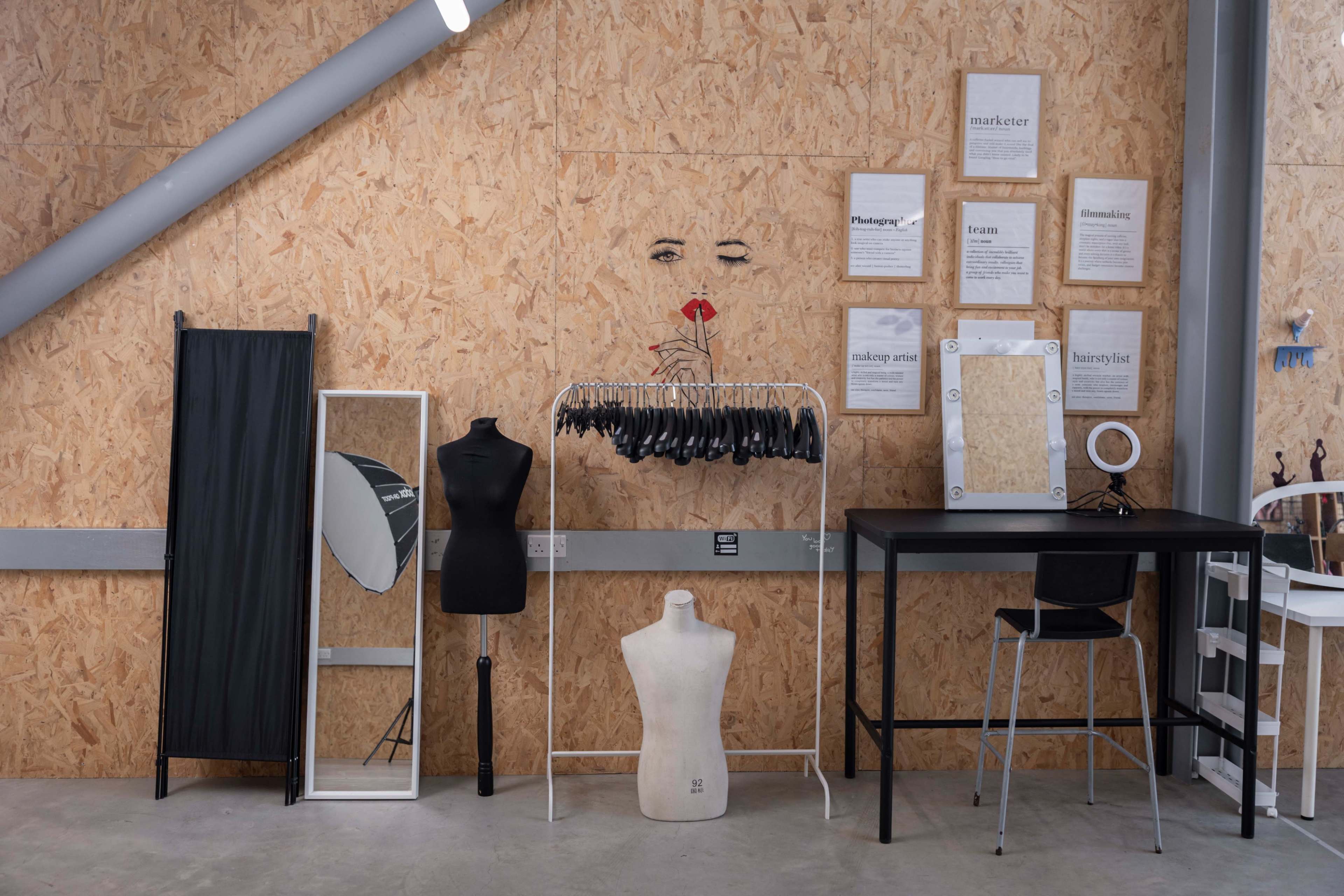 The image shows a workspace with a wooden wall, a clothing rack holding black garments, a mannequin, a full-length mirror, and a small table with a chair.
