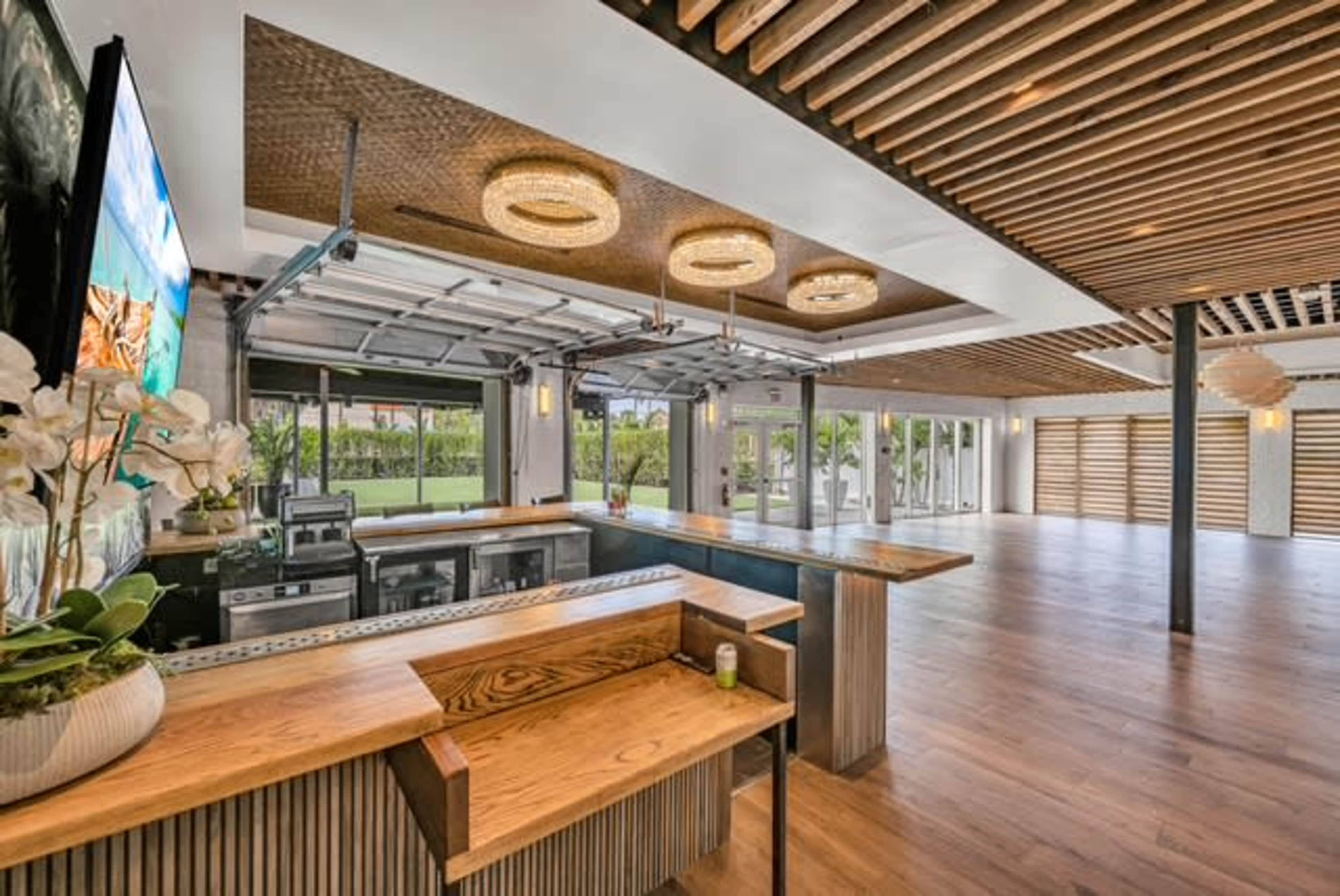 The image shows a contemporary indoor space featuring a bar area with wooden accents, large sliding glass doors, and a spacious floor plan illuminated by overhead lights.
