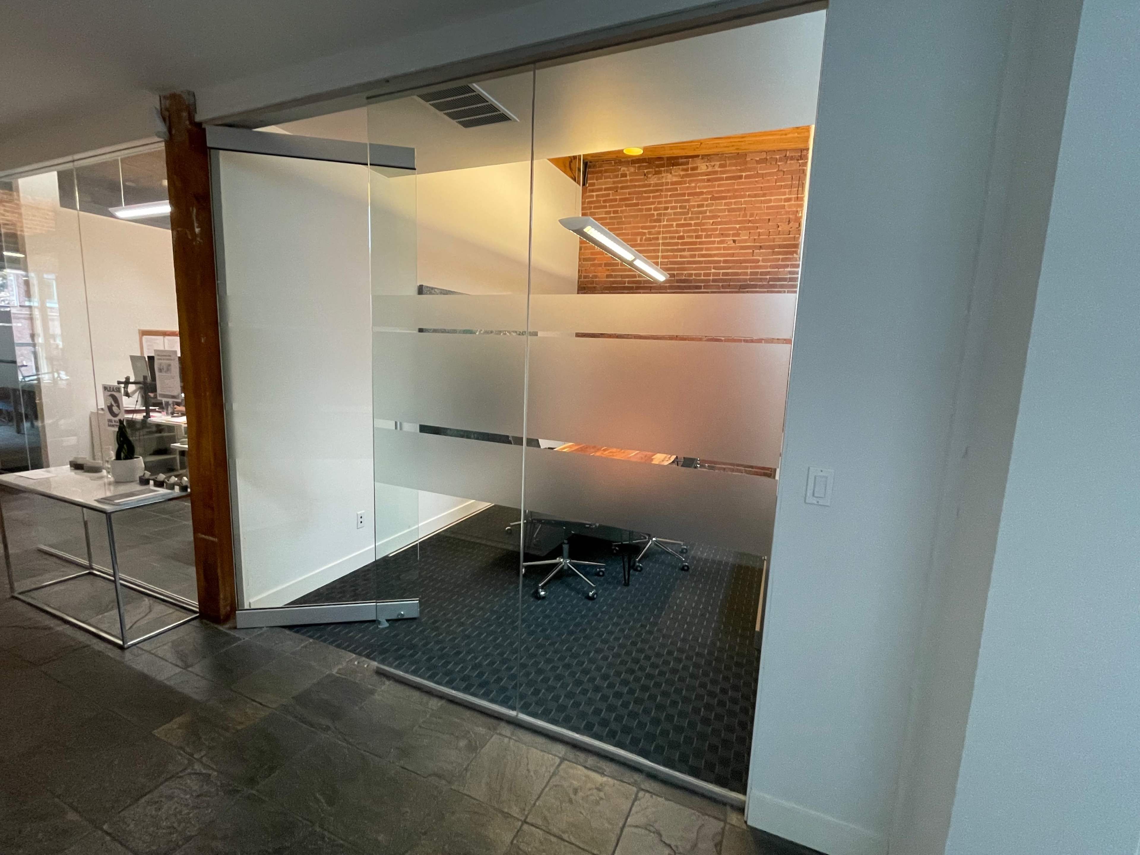 An office space features a glass meeting room with a wooden accent wall and a dark floor mat.