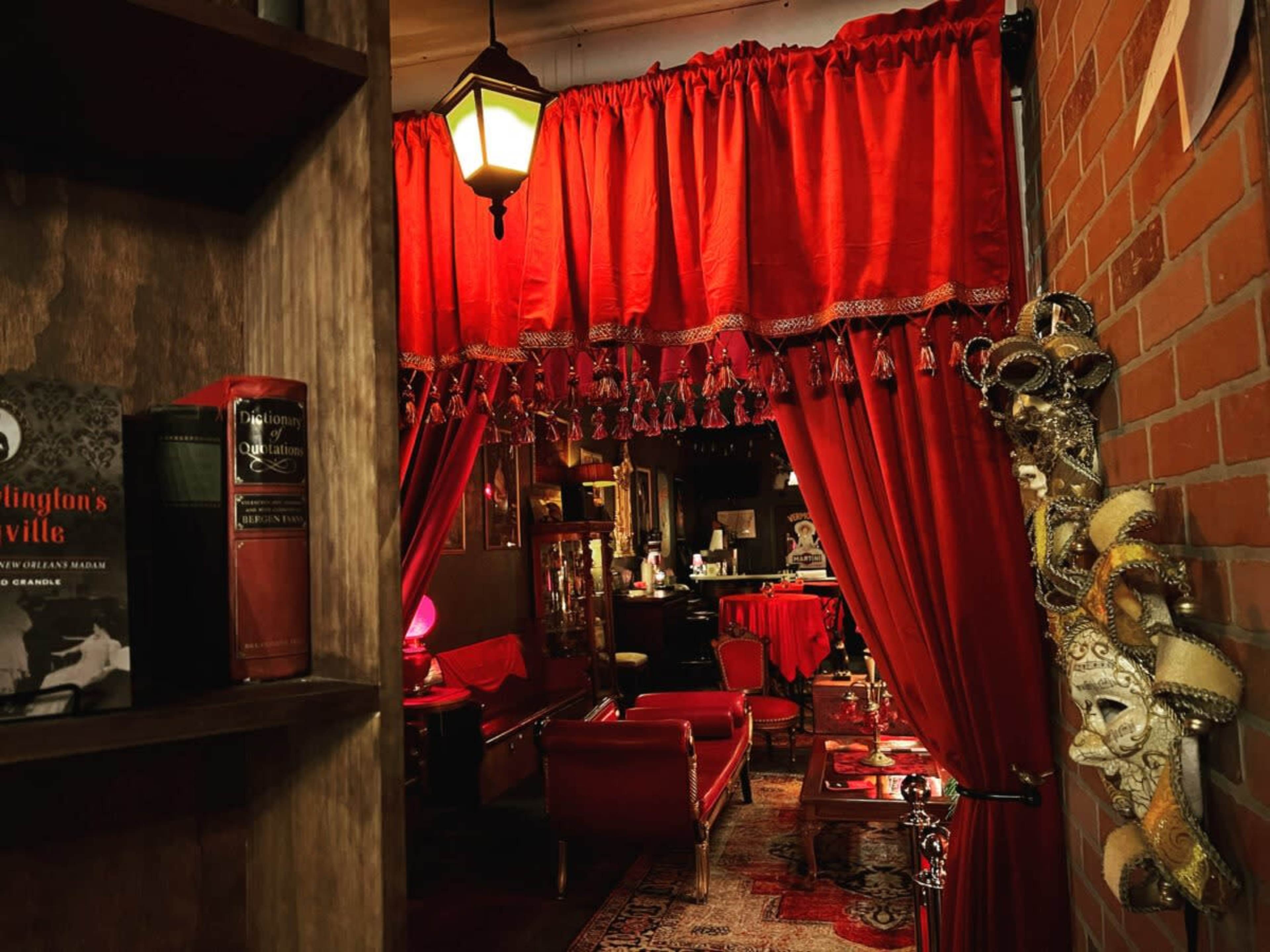 A dimly lit interior room with red curtains, vintage furniture, and decorative masks on a brick wall.