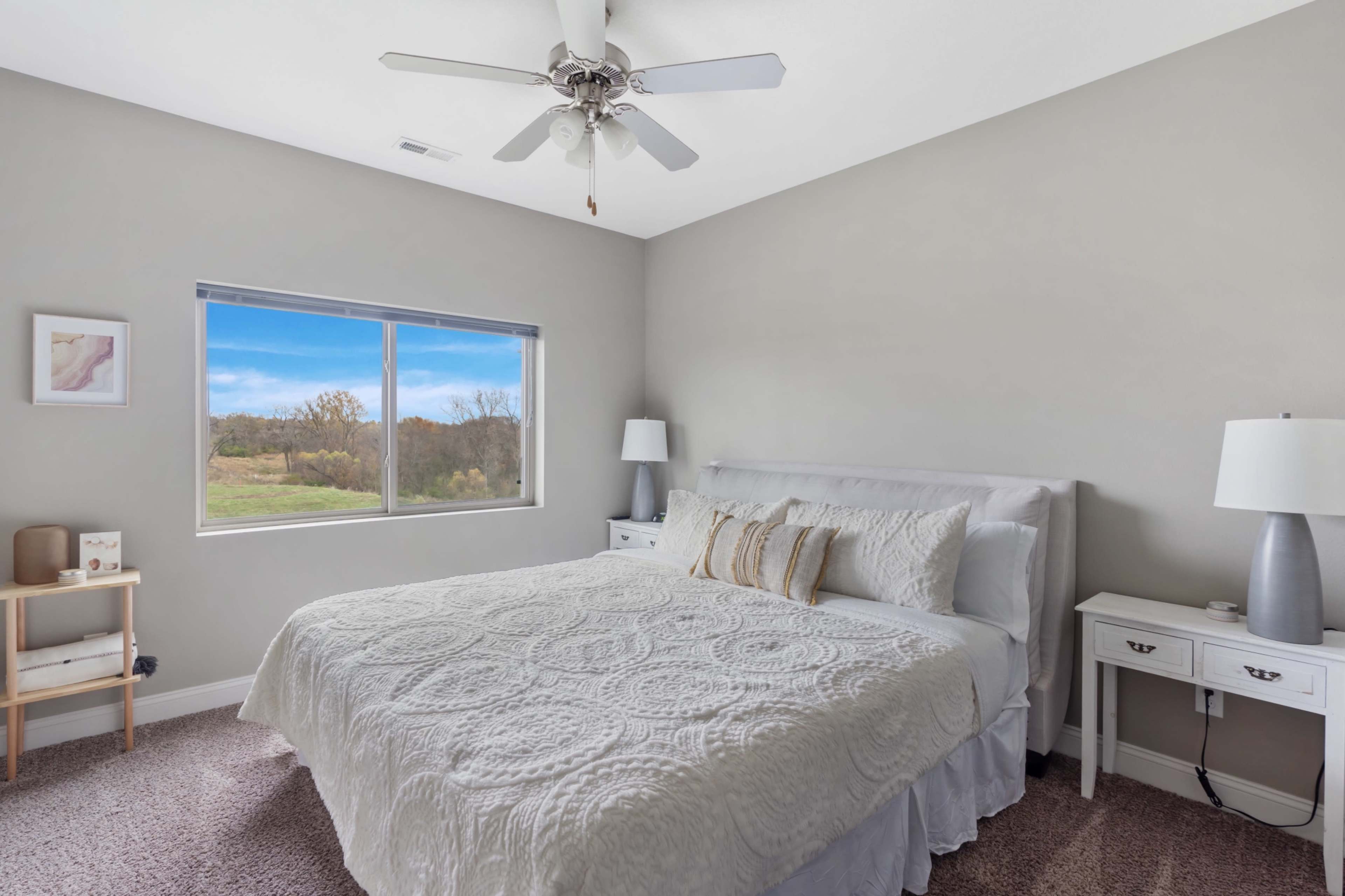 A neatly arranged bedroom featuring a large bed with a white quilt, two bedside tables with lamps, and a window overlooking a grassy landscape.