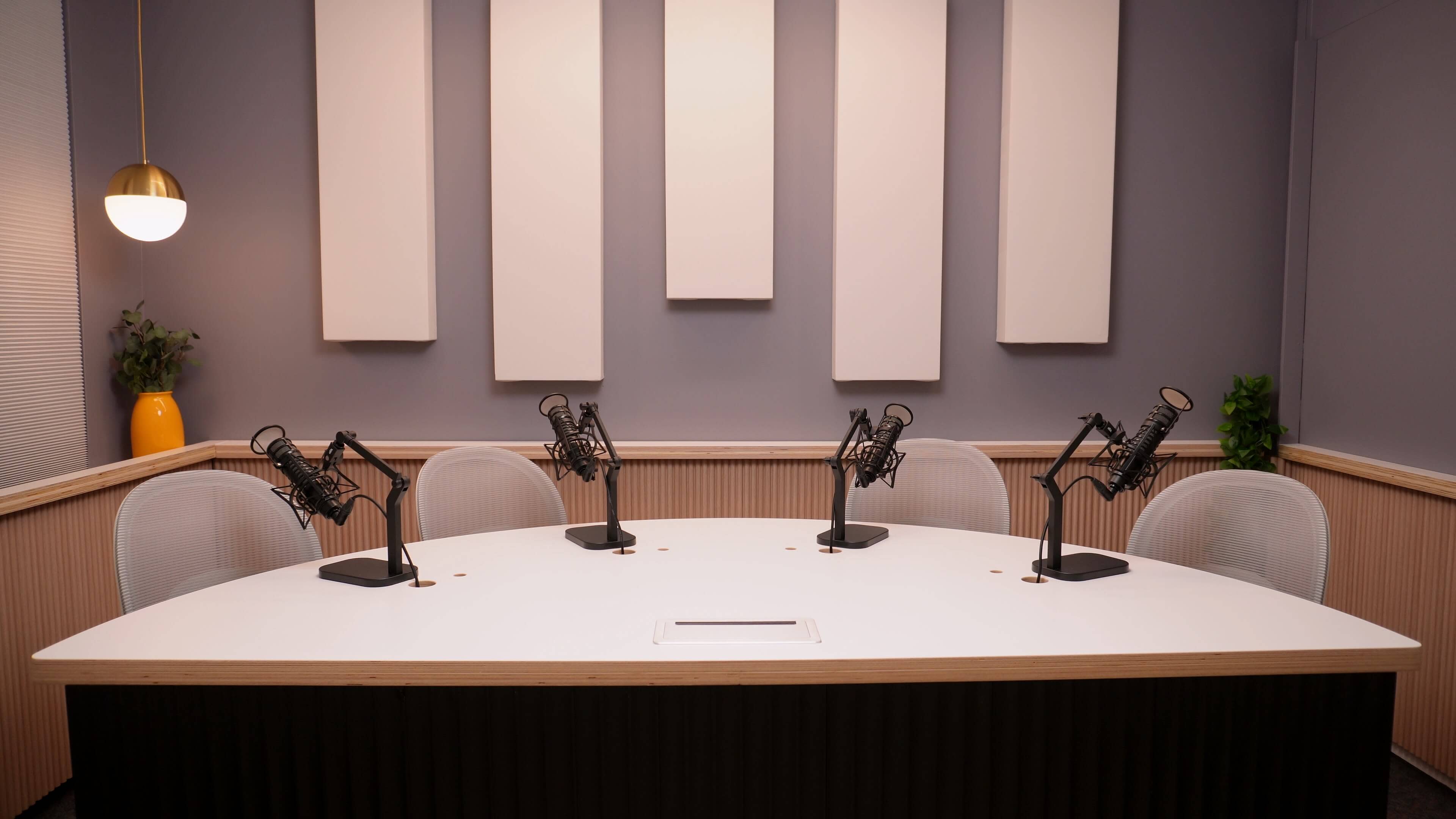 A soundproof podcast studio features a round table with four microphone stands and empty chairs, surrounded by sound-absorbing wall panels.
