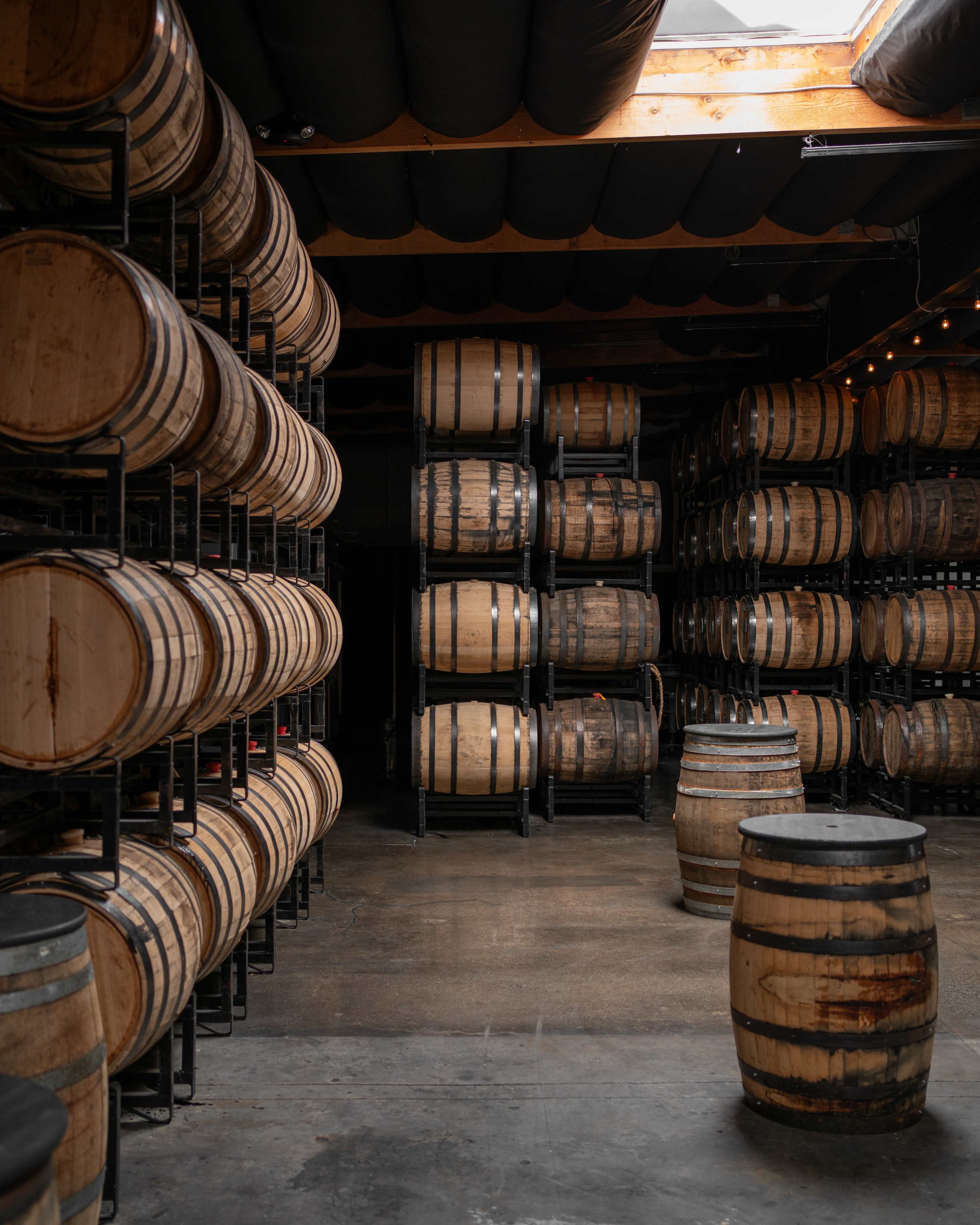 The image depicts a warehouse filled with wooden barrels stacked on metal racks alongside several freestanding barrels.