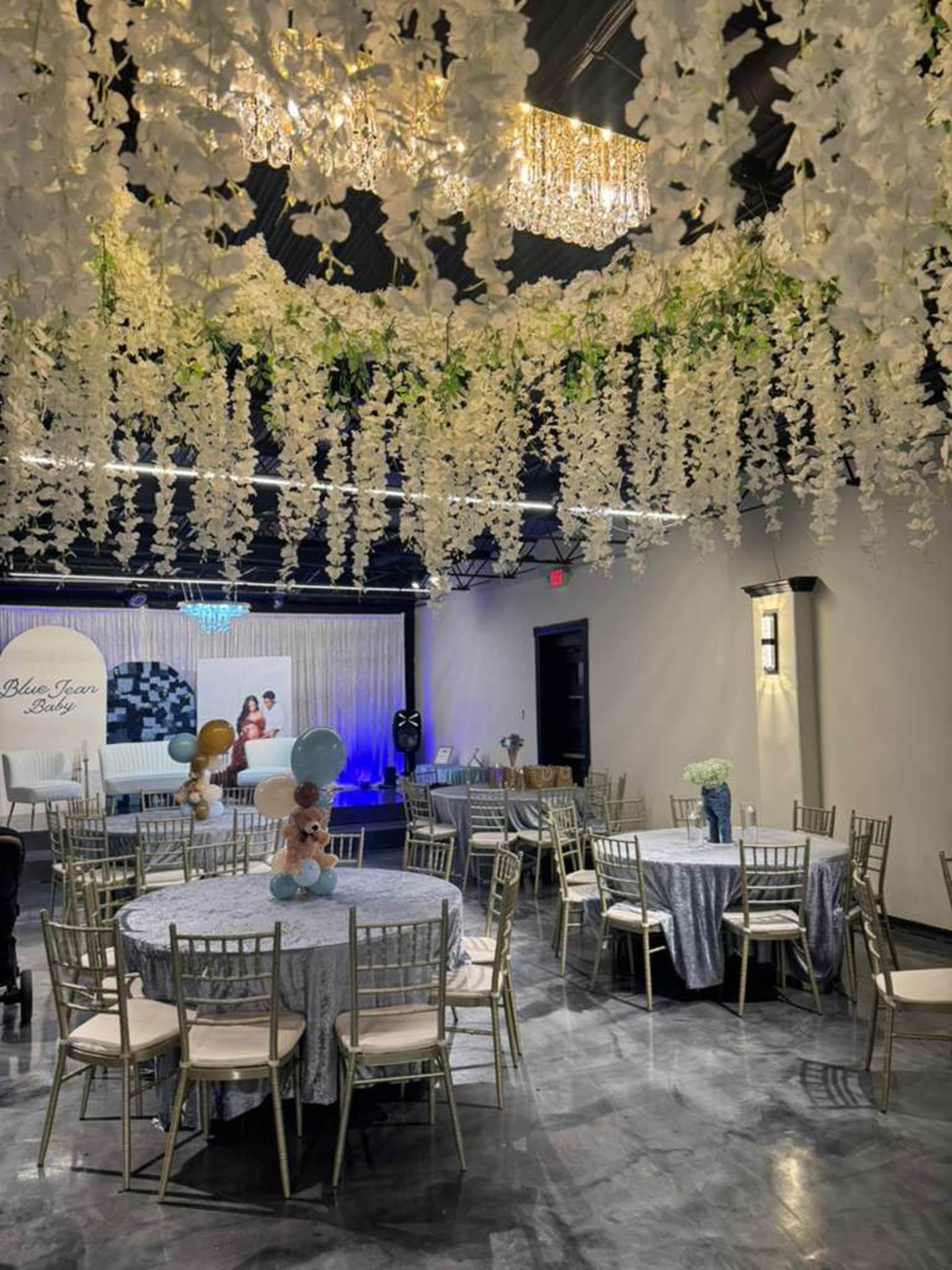 The image shows a decorated event space with a table set for an occasion, featuring hanging floral arrangements and elegant lighting.