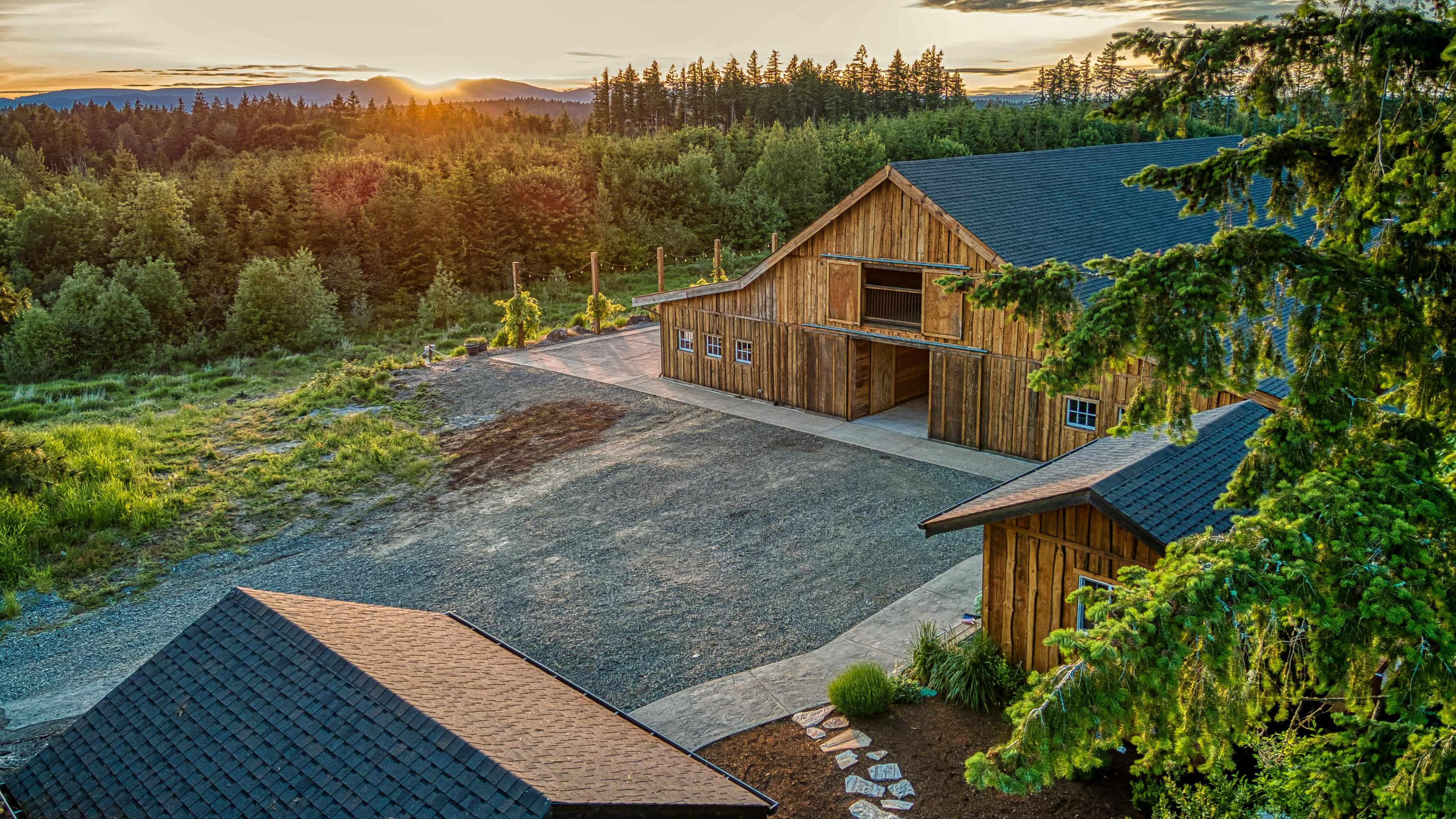 A wooden barn sits on a gravel driveway, surrounded by trees and mountains, as the sun sets in the background.