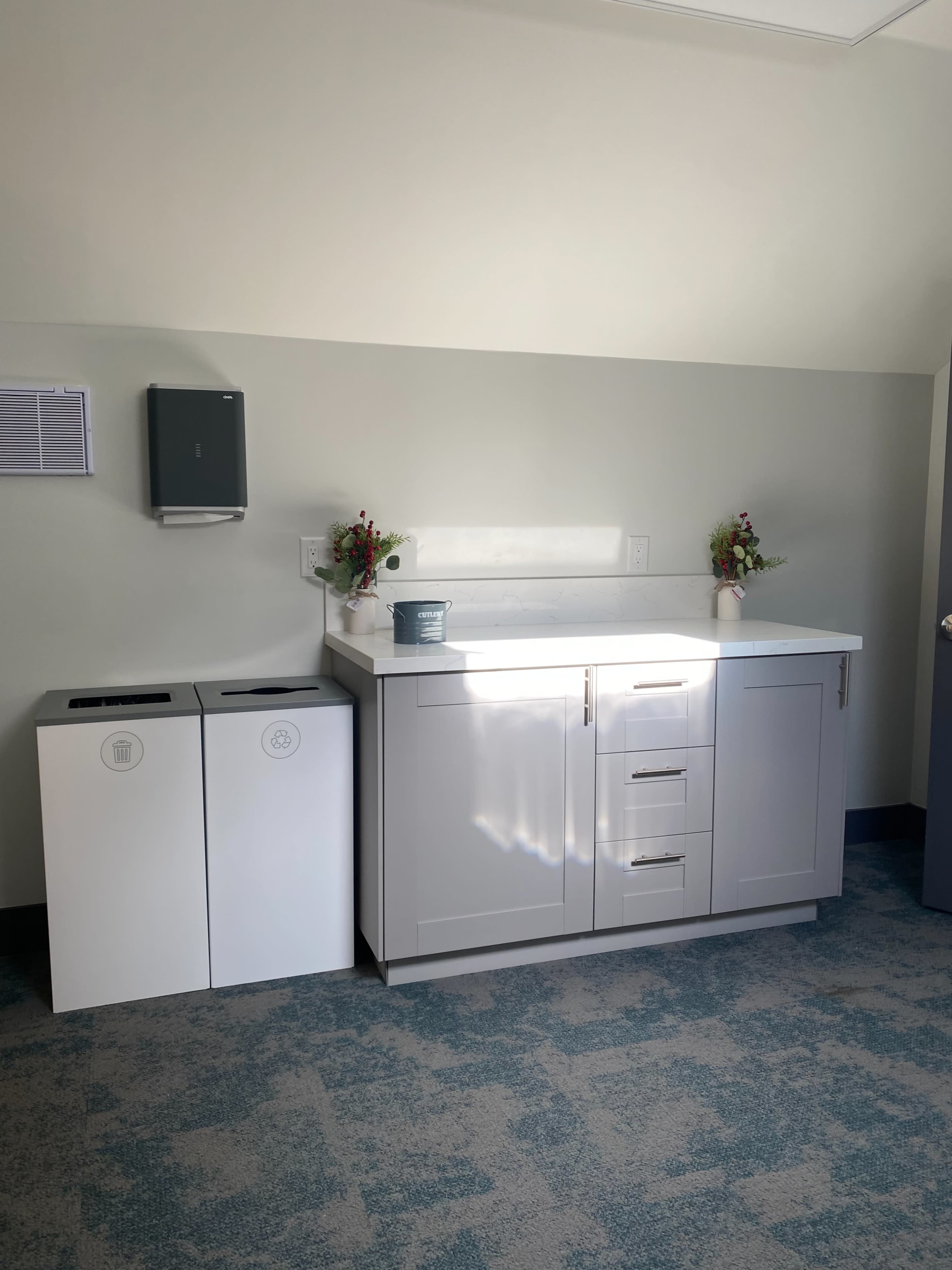 The image shows a clean, modern kitchenette area with a sink, cabinets, and two recycling and trash bins.