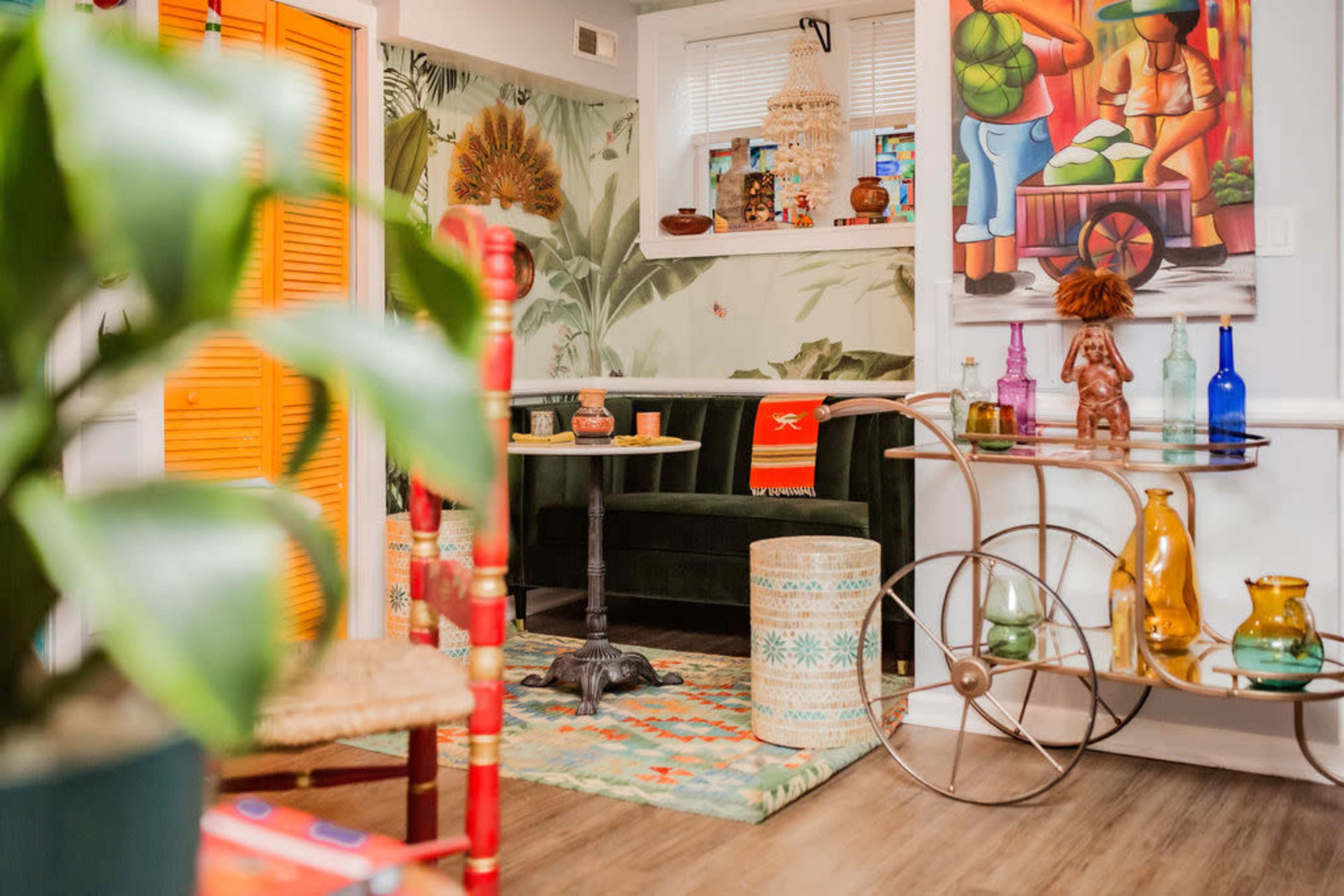 The image shows a colorful interior space with tropical-themed decor, featuring a green sofa, a patterned rug, and various decorative items arranged on a metallic cart.