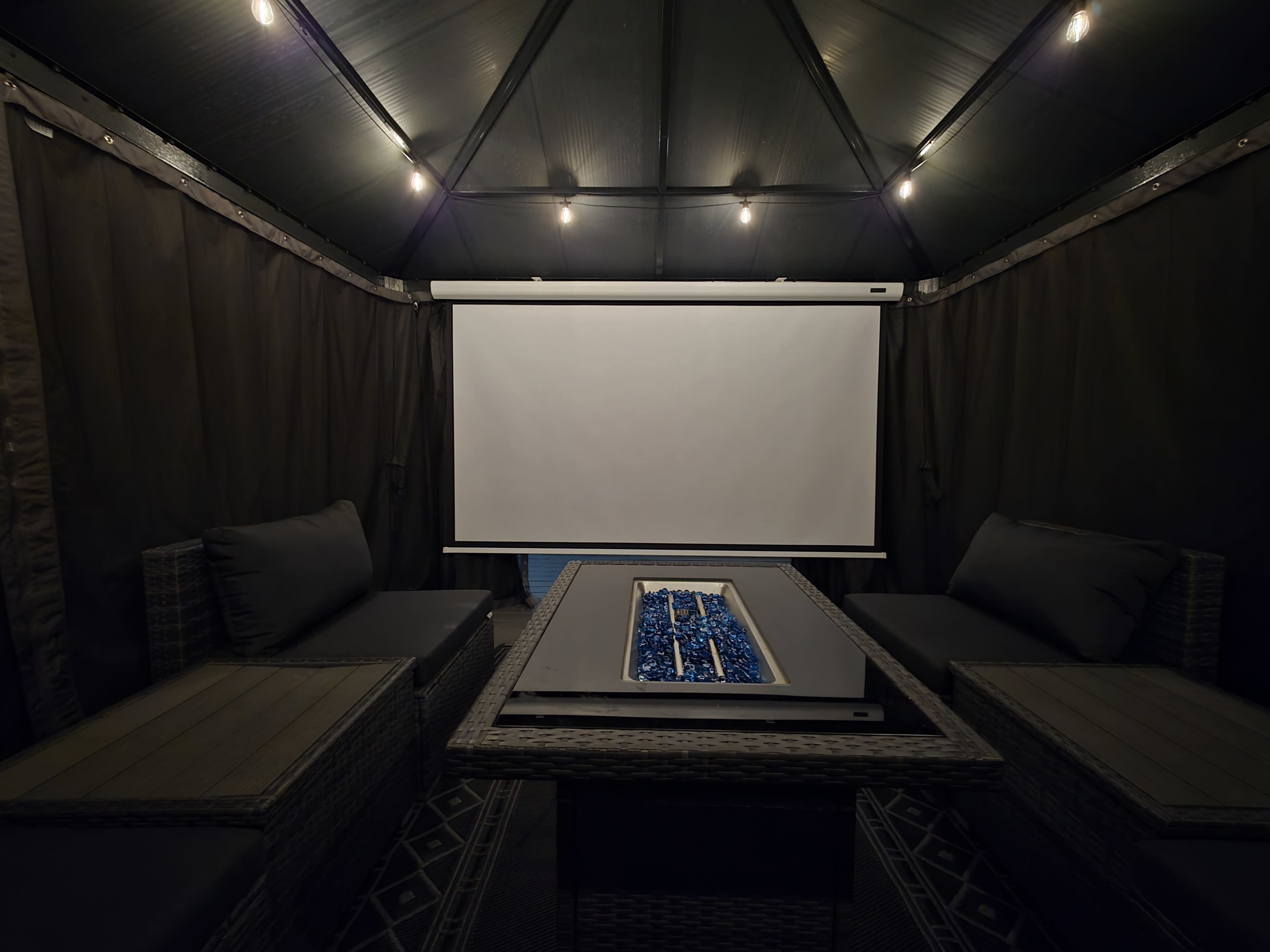 A gazebo is set up with a fire pit table and comfortable seating, featuring a blank projection screen in the center and dim lighting overhead.