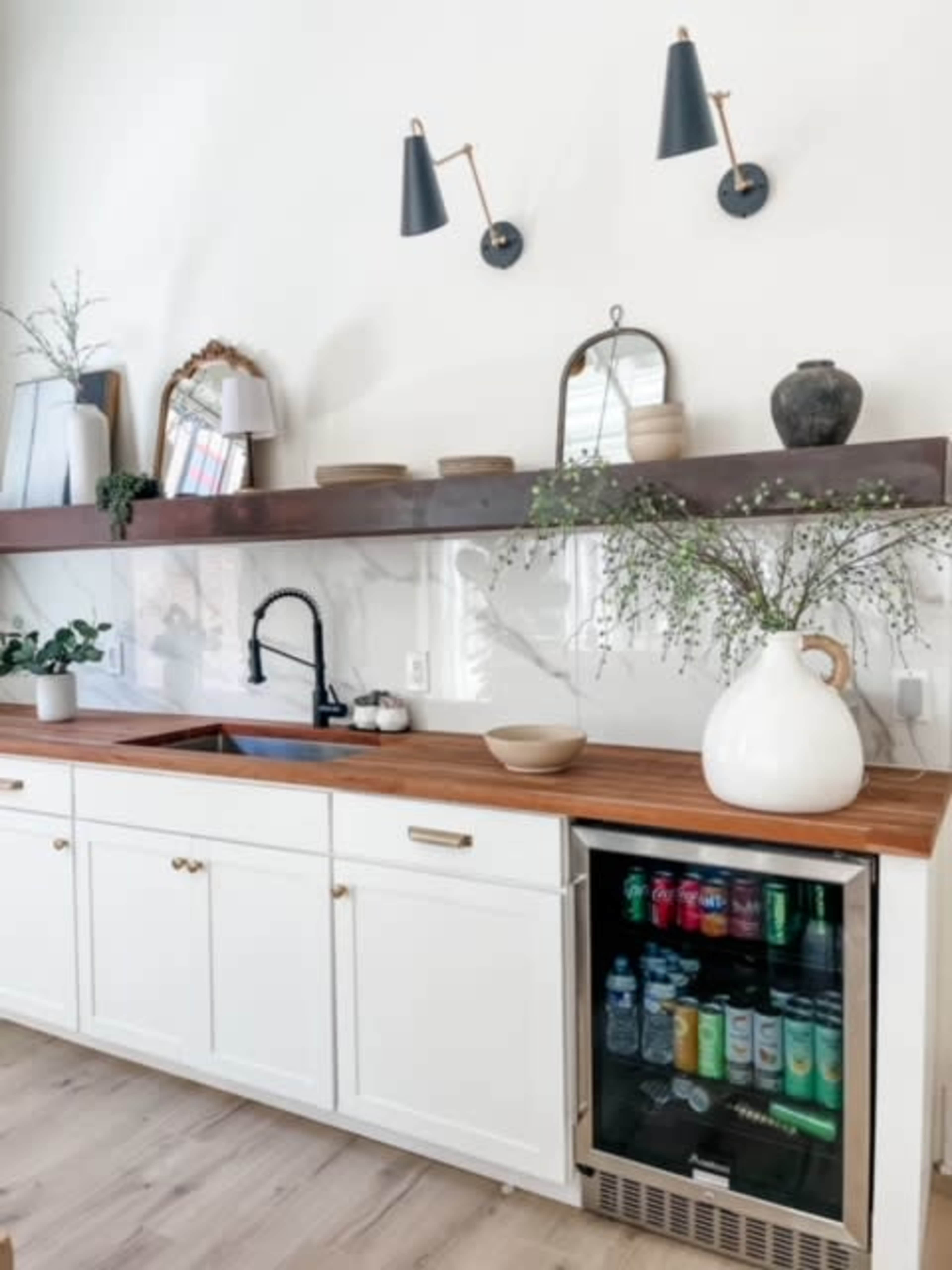 A modern kitchen features white cabinetry, a wooden countertop, wall-mounted sconces, and a mini refrigerator displaying a variety of beverages.