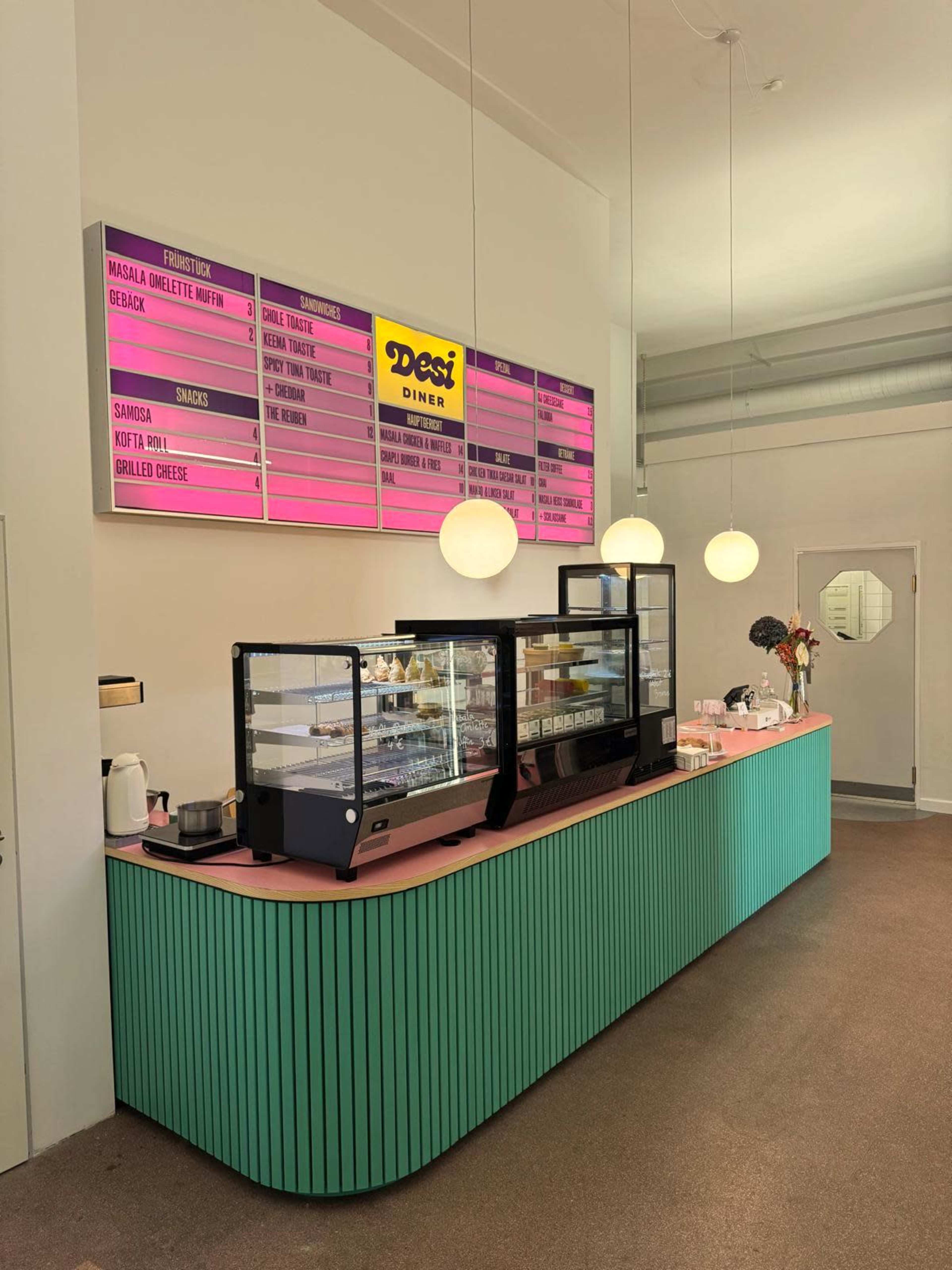 The image shows the interior of a café with a colorful, curved counter displaying pastries and beverages, accompanied by a bright menu board overhead.