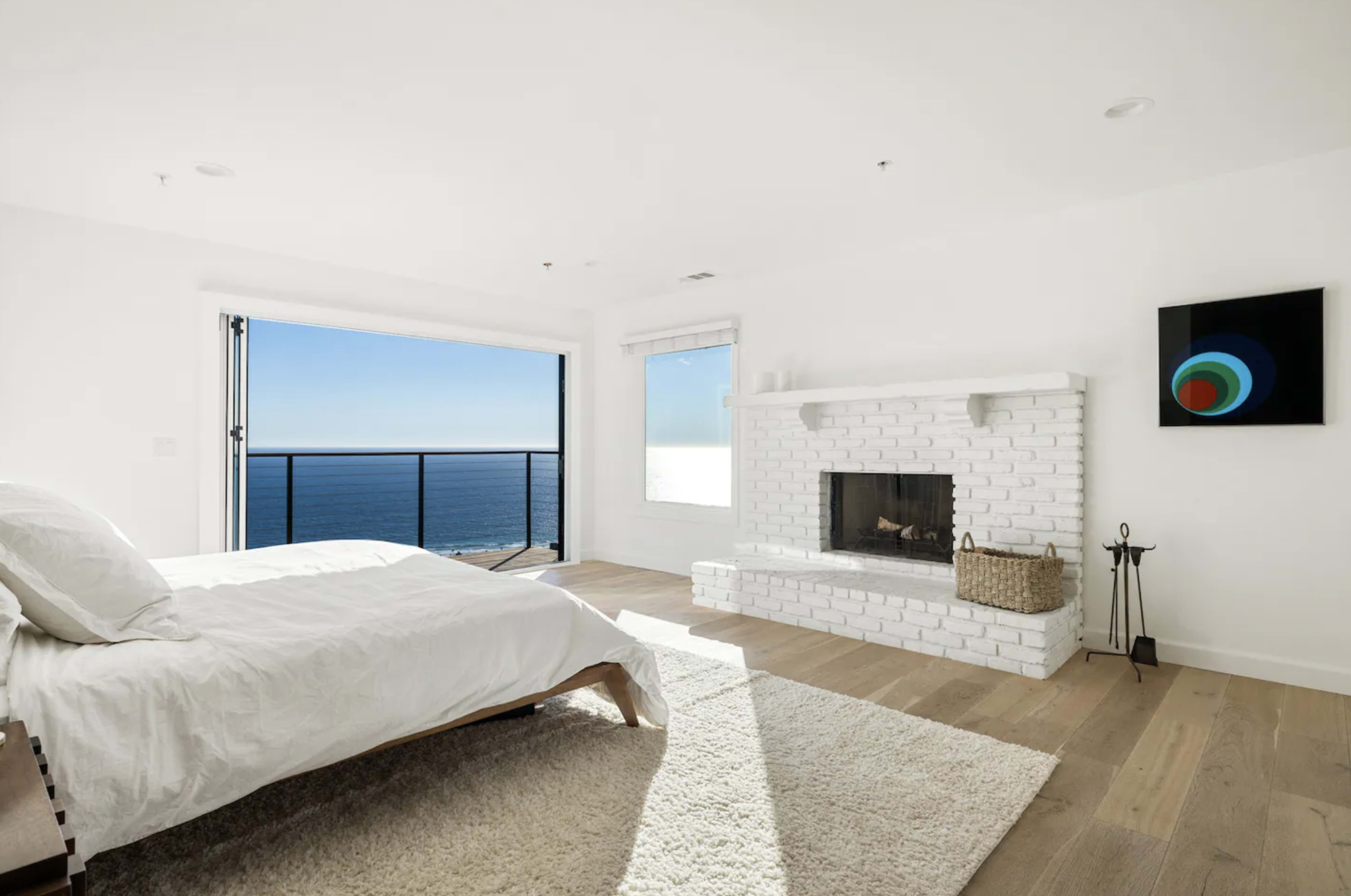 A bright bedroom features a white brick fireplace and a large window overlooking the ocean.