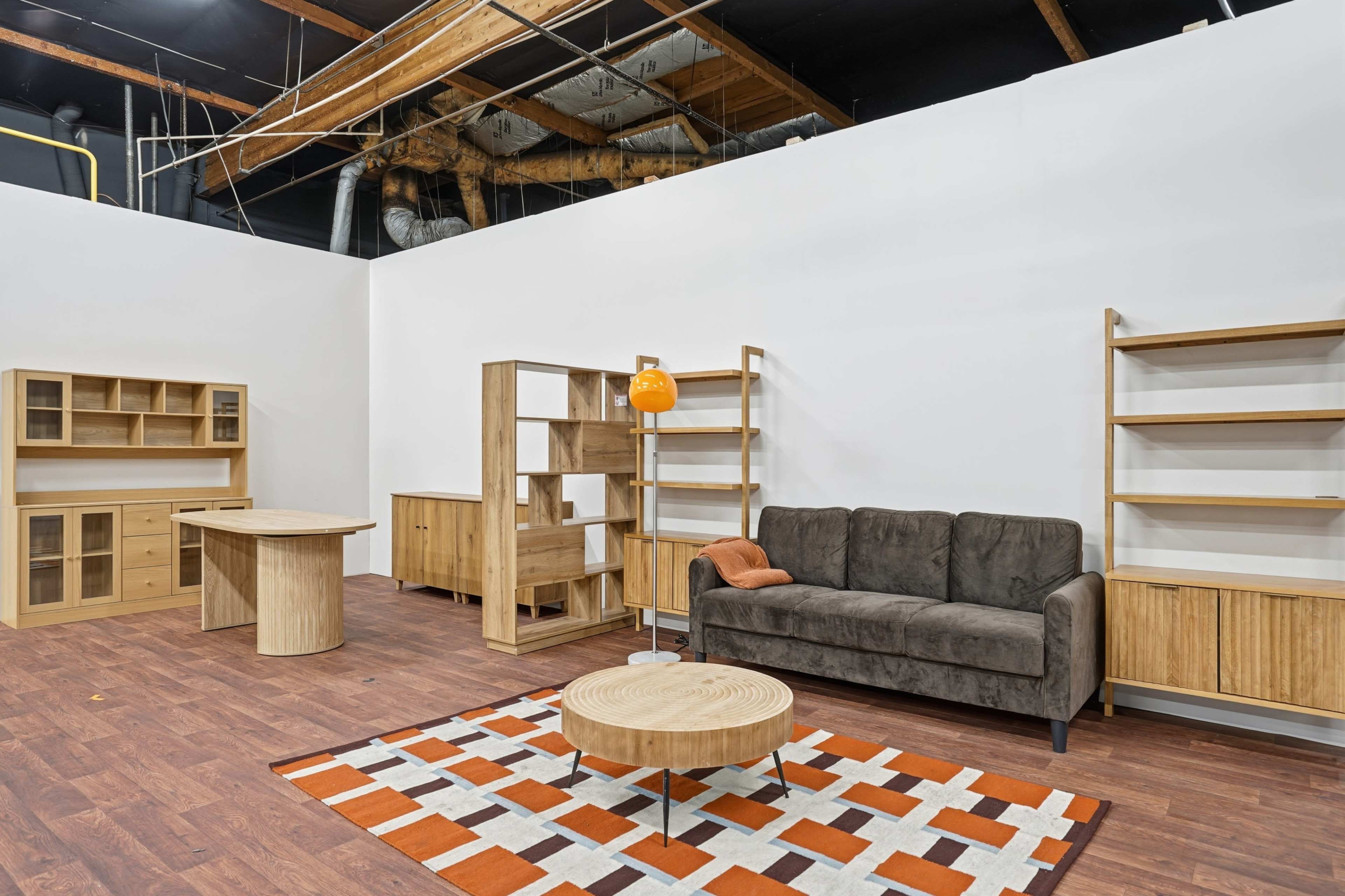 The image shows a modern interior space featuring wooden furniture, including a sofa, a coffee table, bookshelves, and a desk, against a plain white wall.