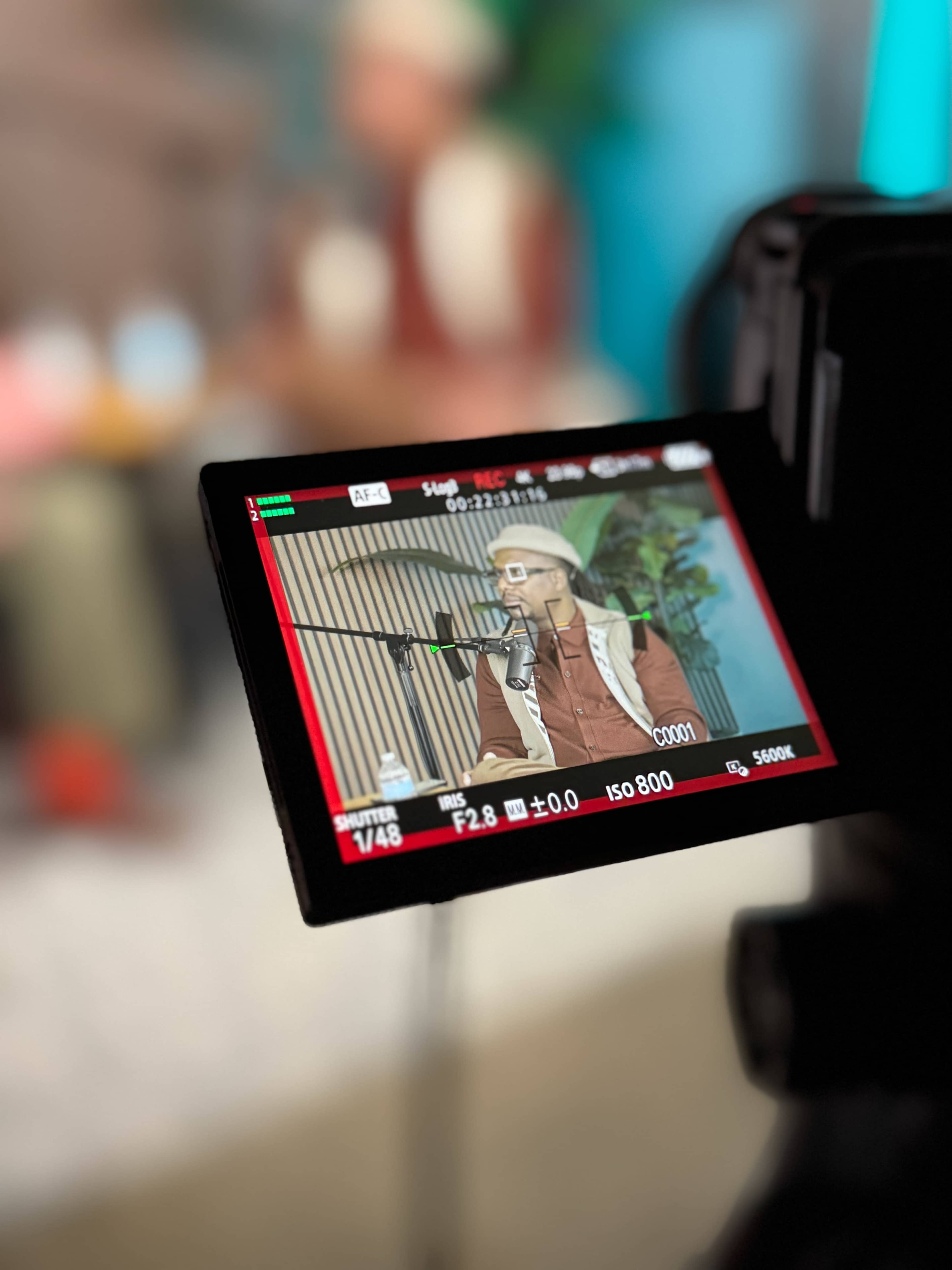 A camera captures an individual speaking in a studio setting, with a blurred background and equipment visible.