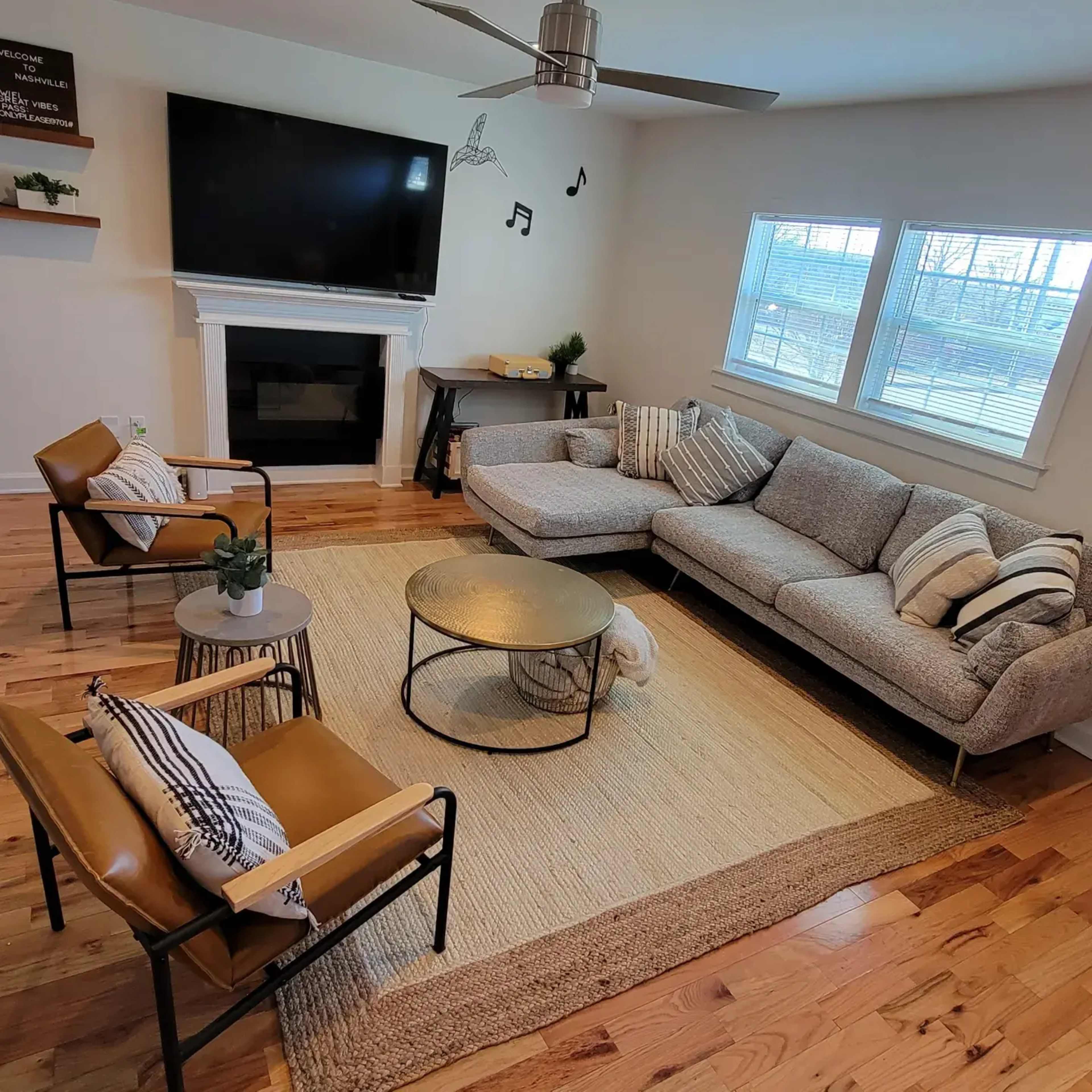 A modern living room features a large sectional sofa, a round coffee table, two armchairs, and a television above a fireplace, all arranged on a wooden floor with a textured area rug.