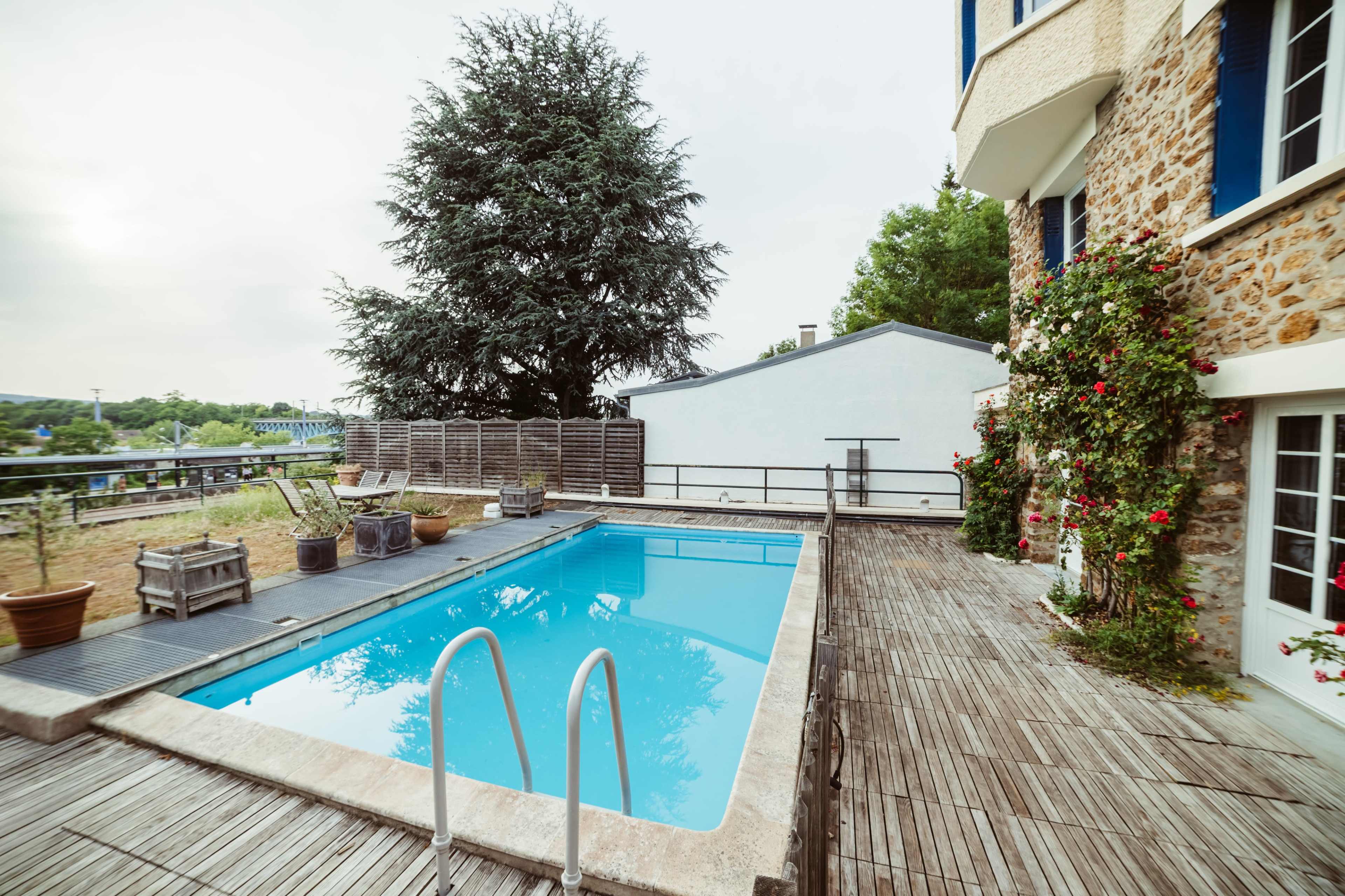 The image shows a rectangular swimming pool surrounded by a wooden deck, with potted plants and a large tree in the background.