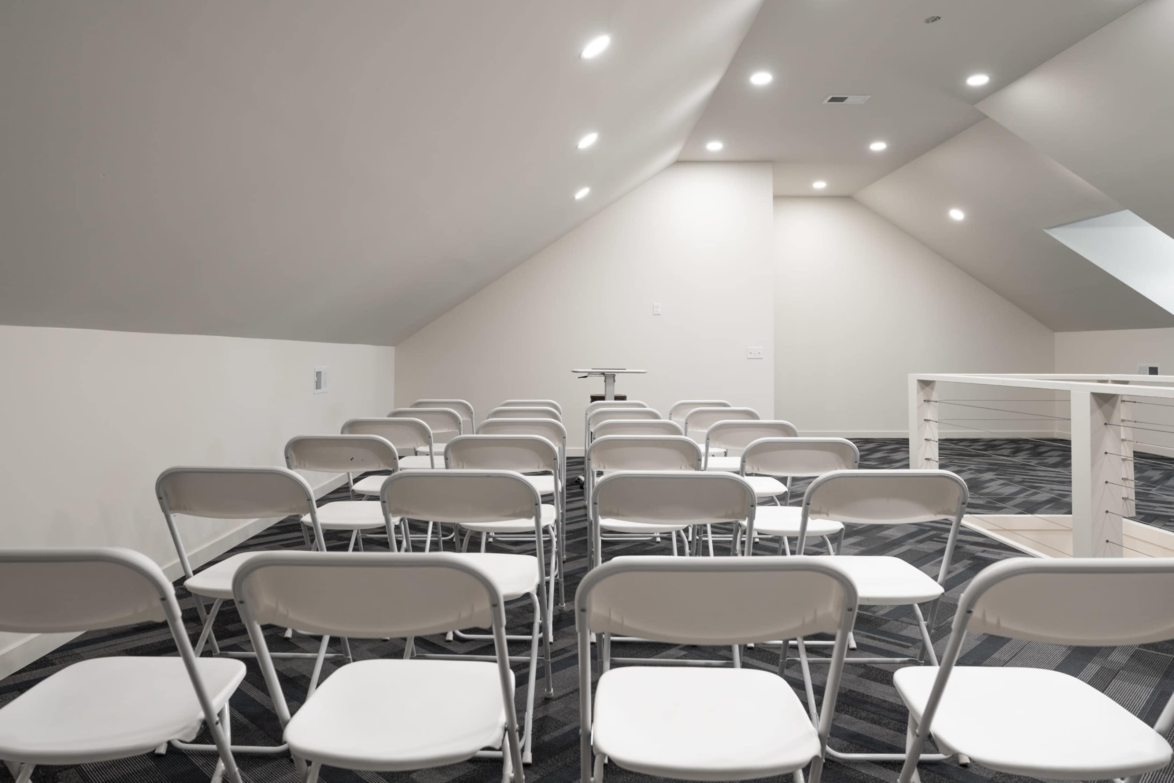 A room with sloped ceilings arranged with several rows of white folding chairs facing a central table.
